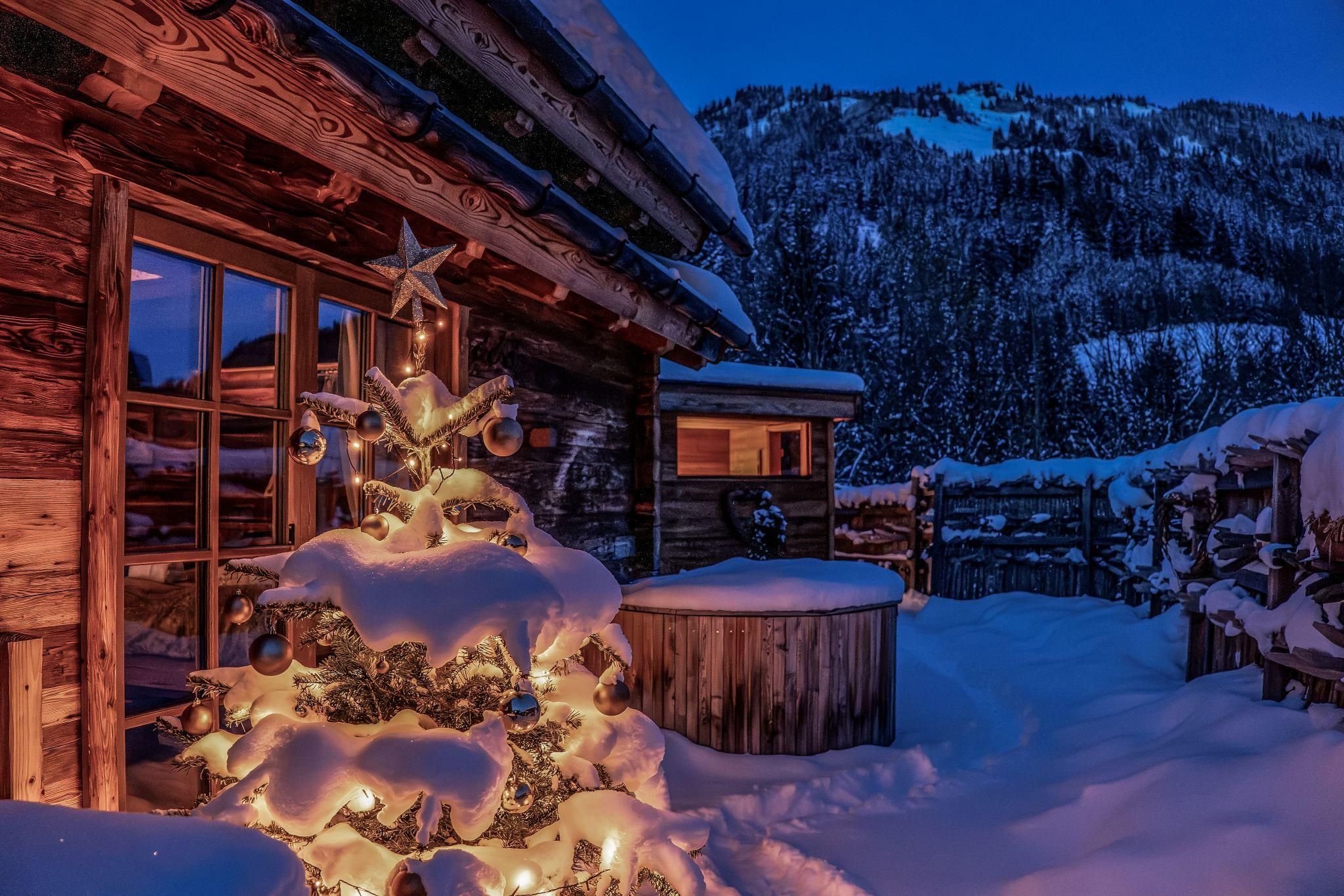 A wintry landscape with a Christmas tree adorned with lights. In the background, there are snow-covered mountains and a chalet.