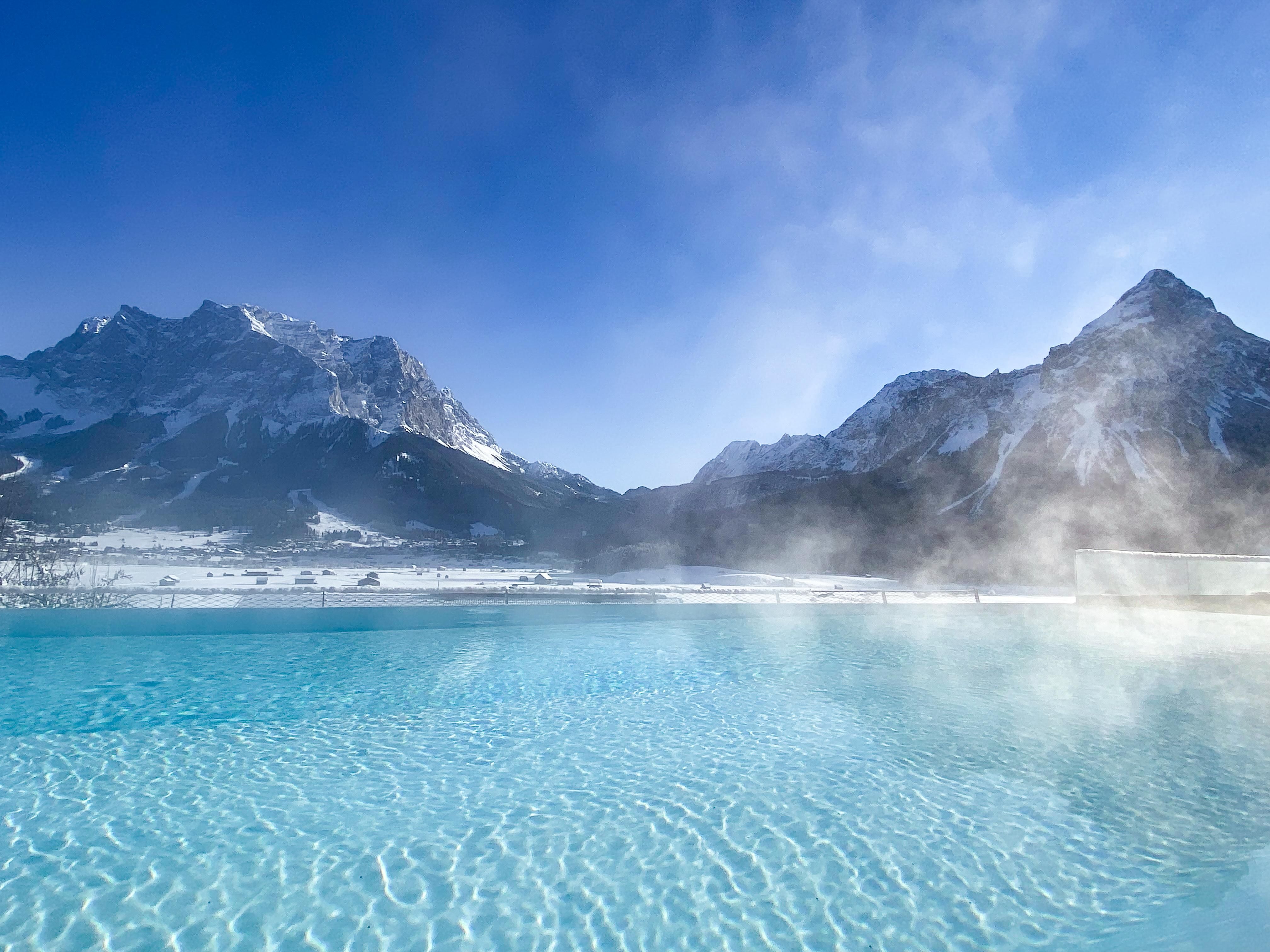 A beautiful view of sparkling, turquoise water in front of snow-covered mountains. The sky is clear and blue, with a hint of mist over the water's surface.
