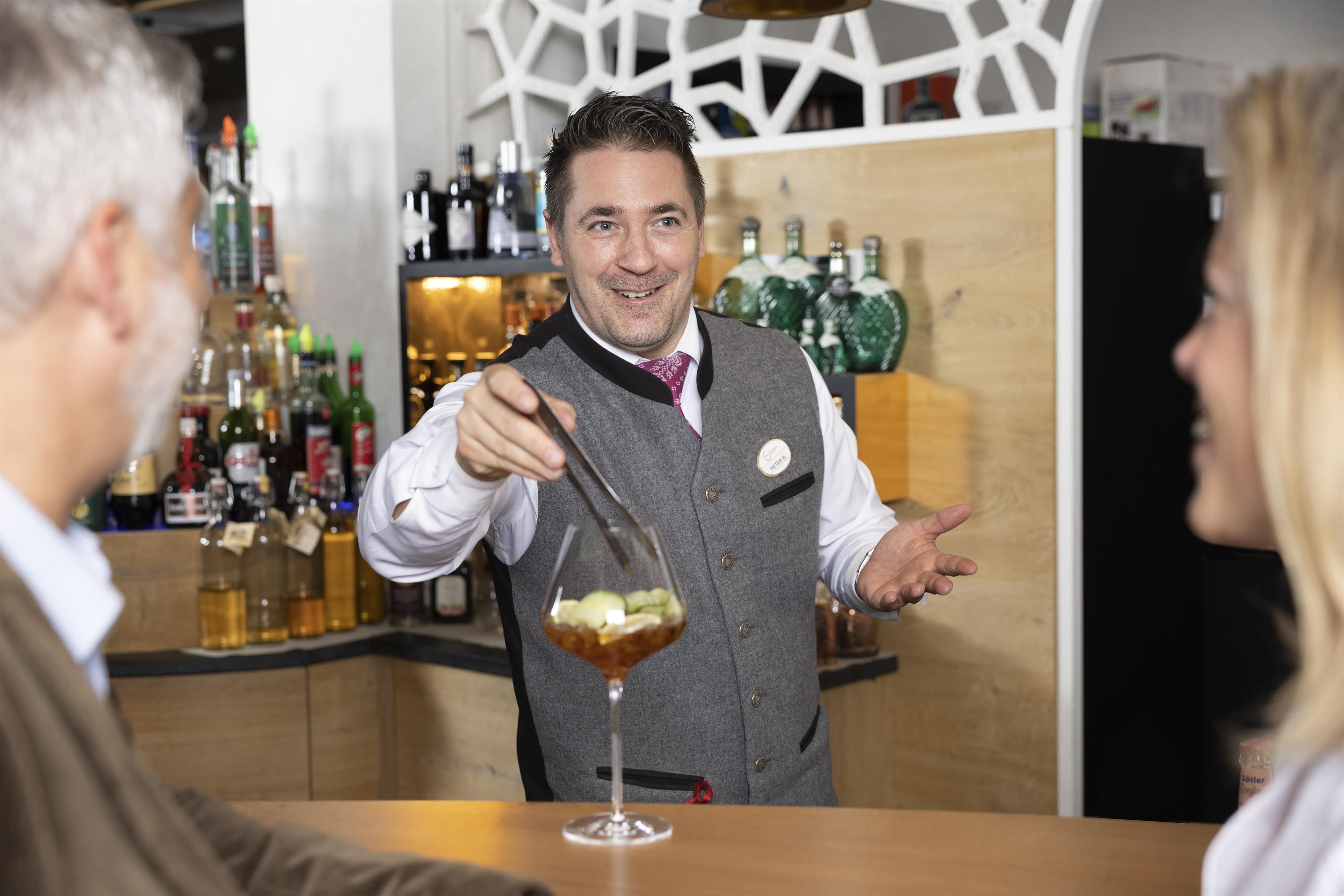 A bartender serves a cocktail to guests. The bartender smiles kindly and presents the drink with an inviting gesture.
