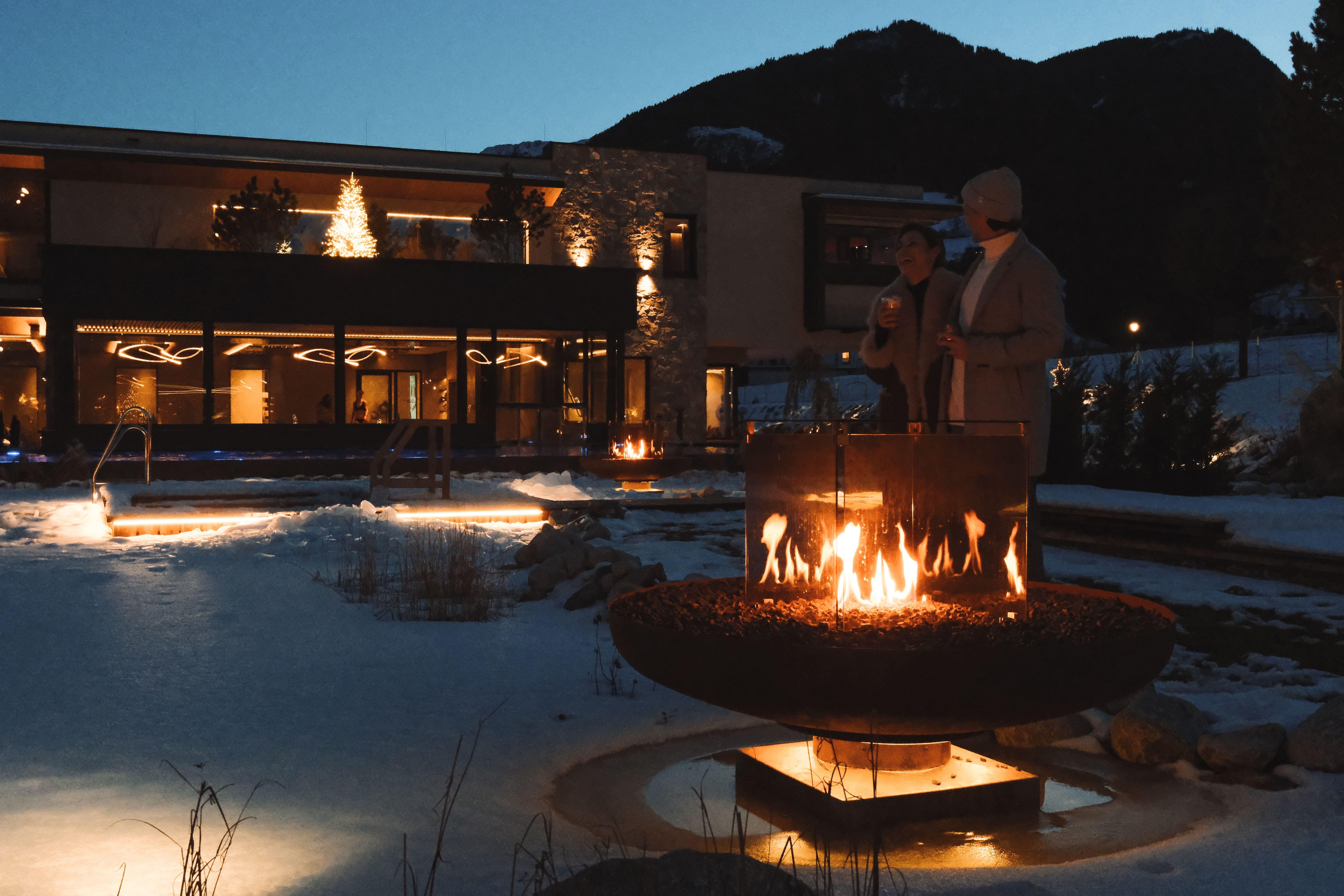 A cozy winter landscape with a fire in a large metal ring. In the background, a stylish building can be seen with Christmas lights and a snow-covered environment.