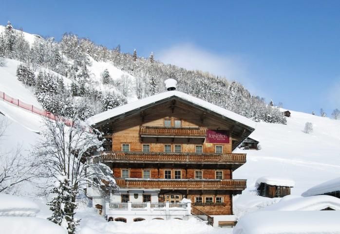 A traditional wooden house in a snowy winter landscape. In the background, snow-covered mountains and trees can be seen.