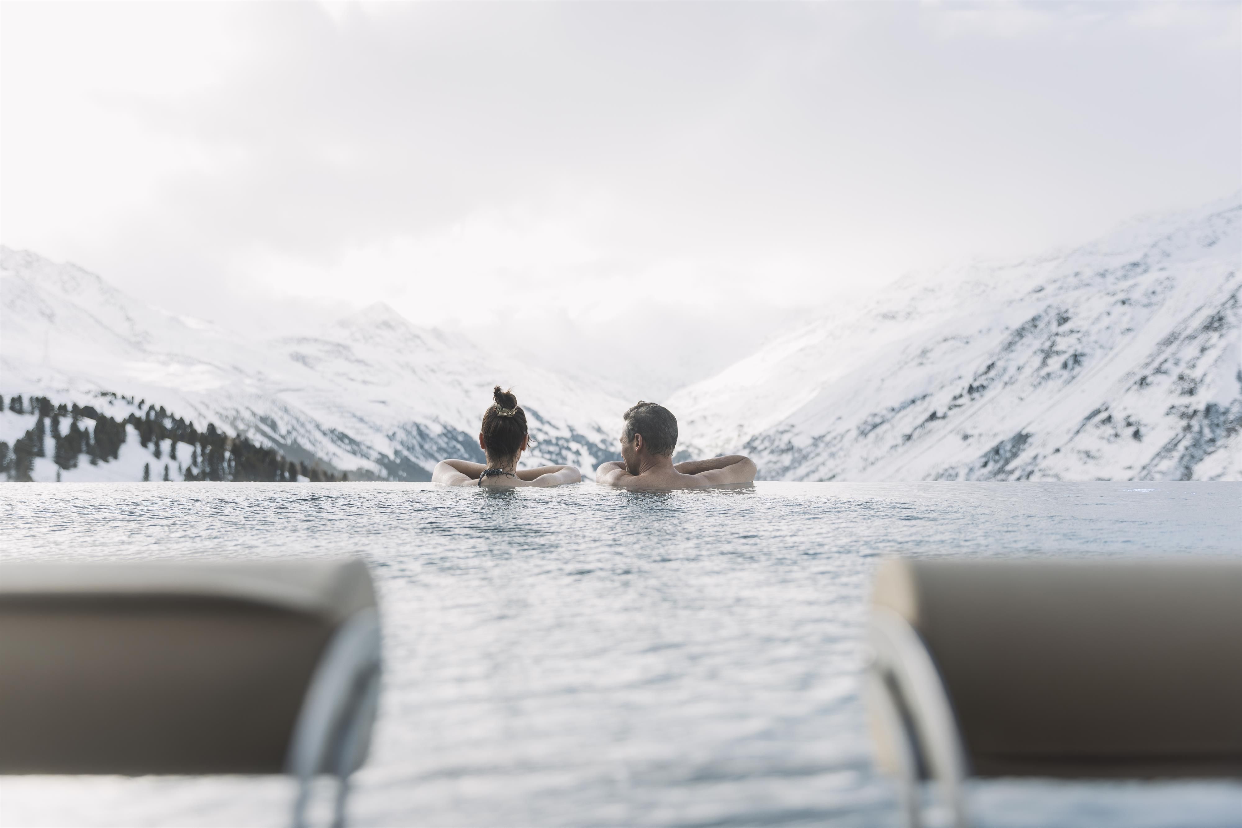 A couple relaxes in the infinity pool with a view of snow-covered mountains. The calm atmosphere and the snowy landscape create a relaxing mood.