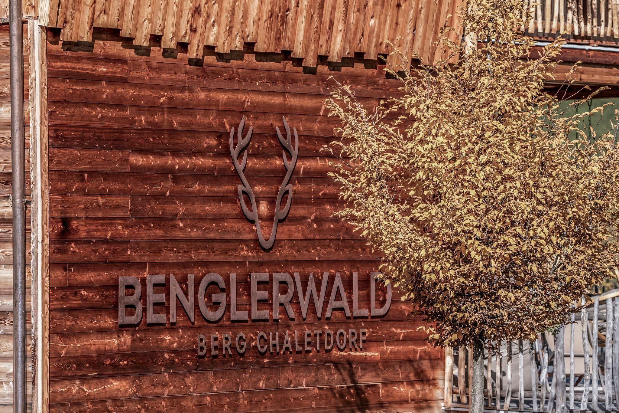 A wooden sign with the inscription "Benglerwald Mountain Chalet Village" and a deer antler symbol. In the foreground stands a tree with yellow leaves.