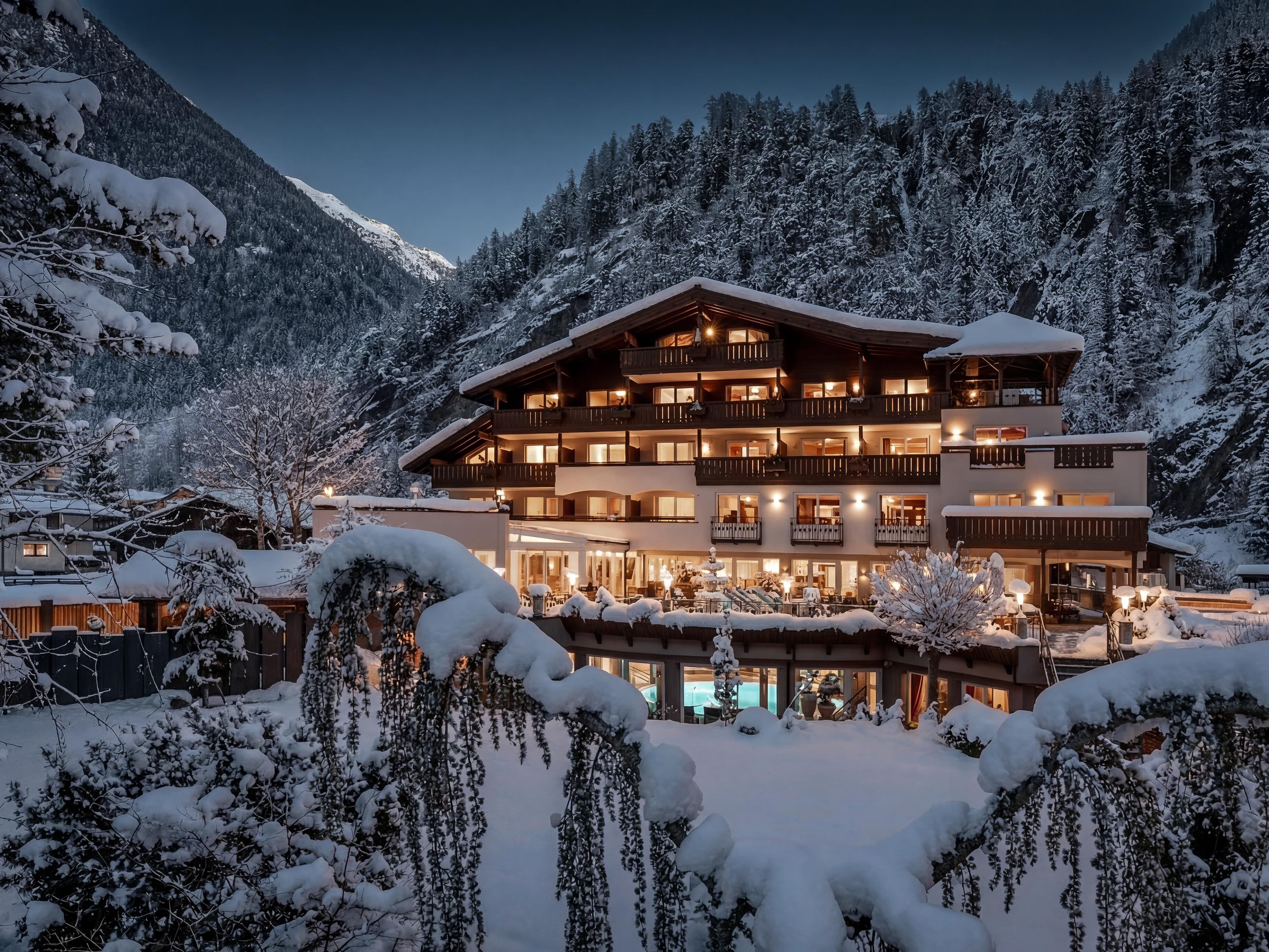 A charming hotel in winter, surrounded by snow-covered mountains. The facade is illuminated and the surroundings are enveloped in a calm, wintry atmosphere.