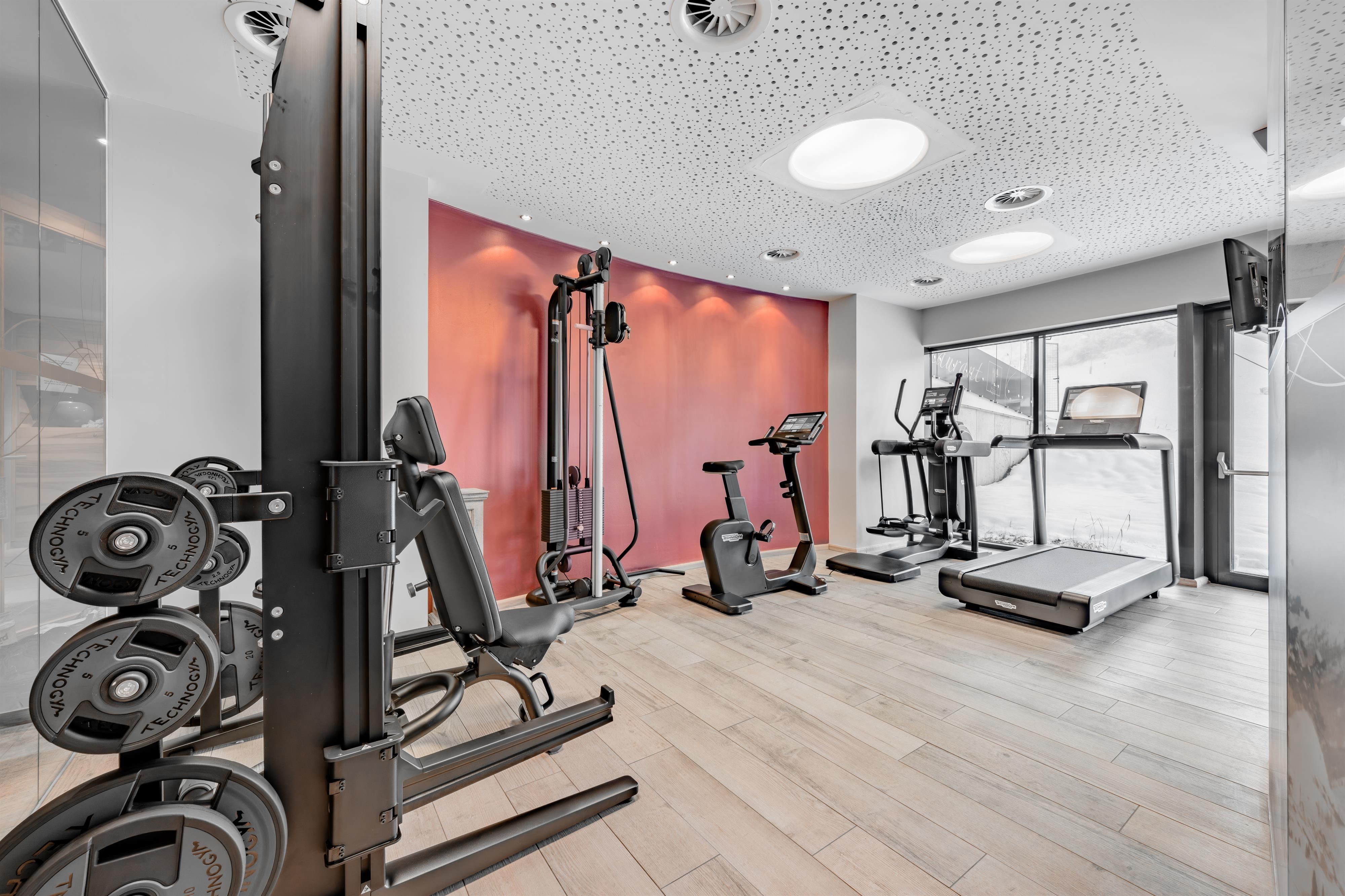 A modern gym with several training machines. The room is bright and well-equipped with wooden flooring.