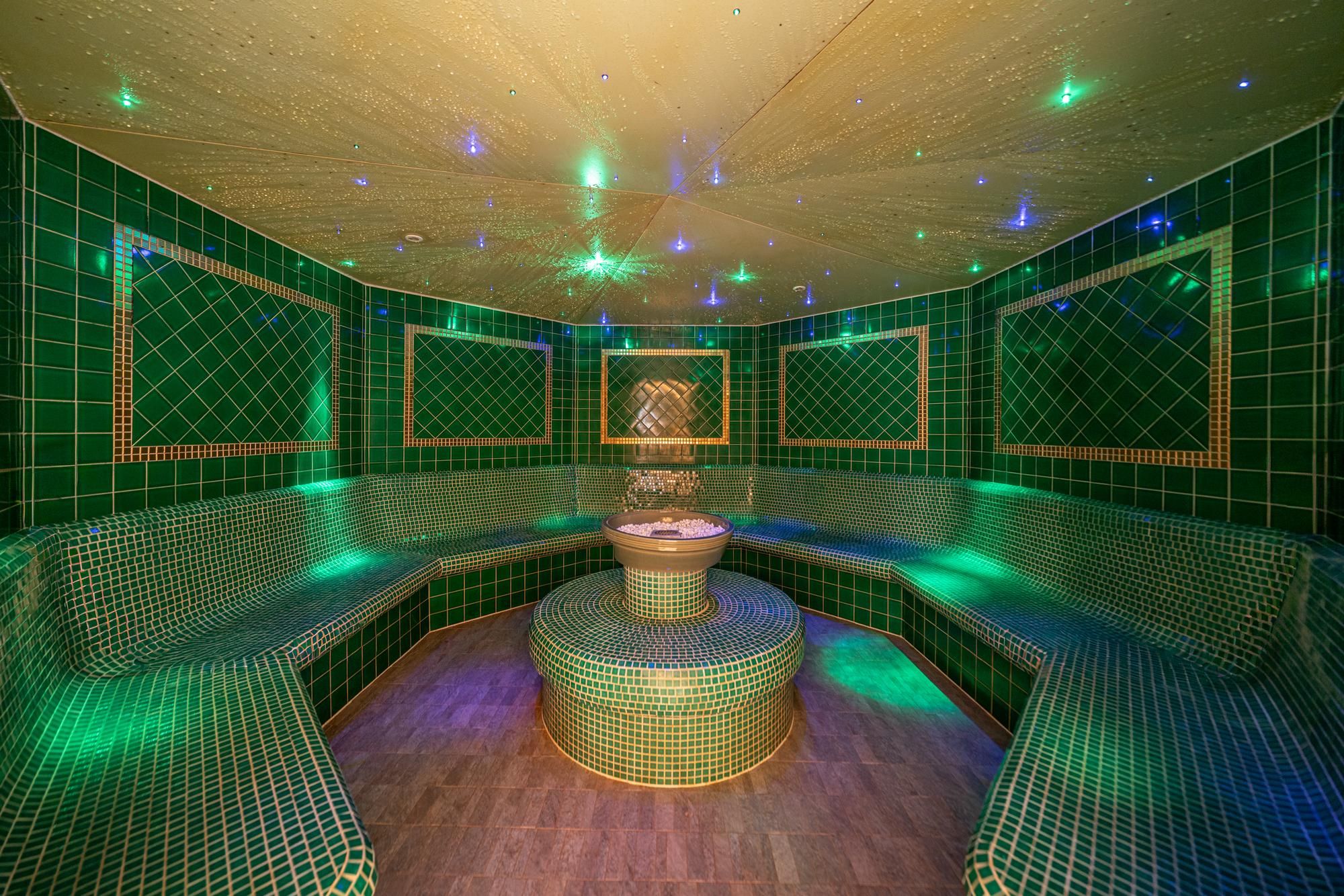 A luxurious steam bath with green tiles and soft lighting effects. A round seating area in the center creates a relaxing atmosphere.