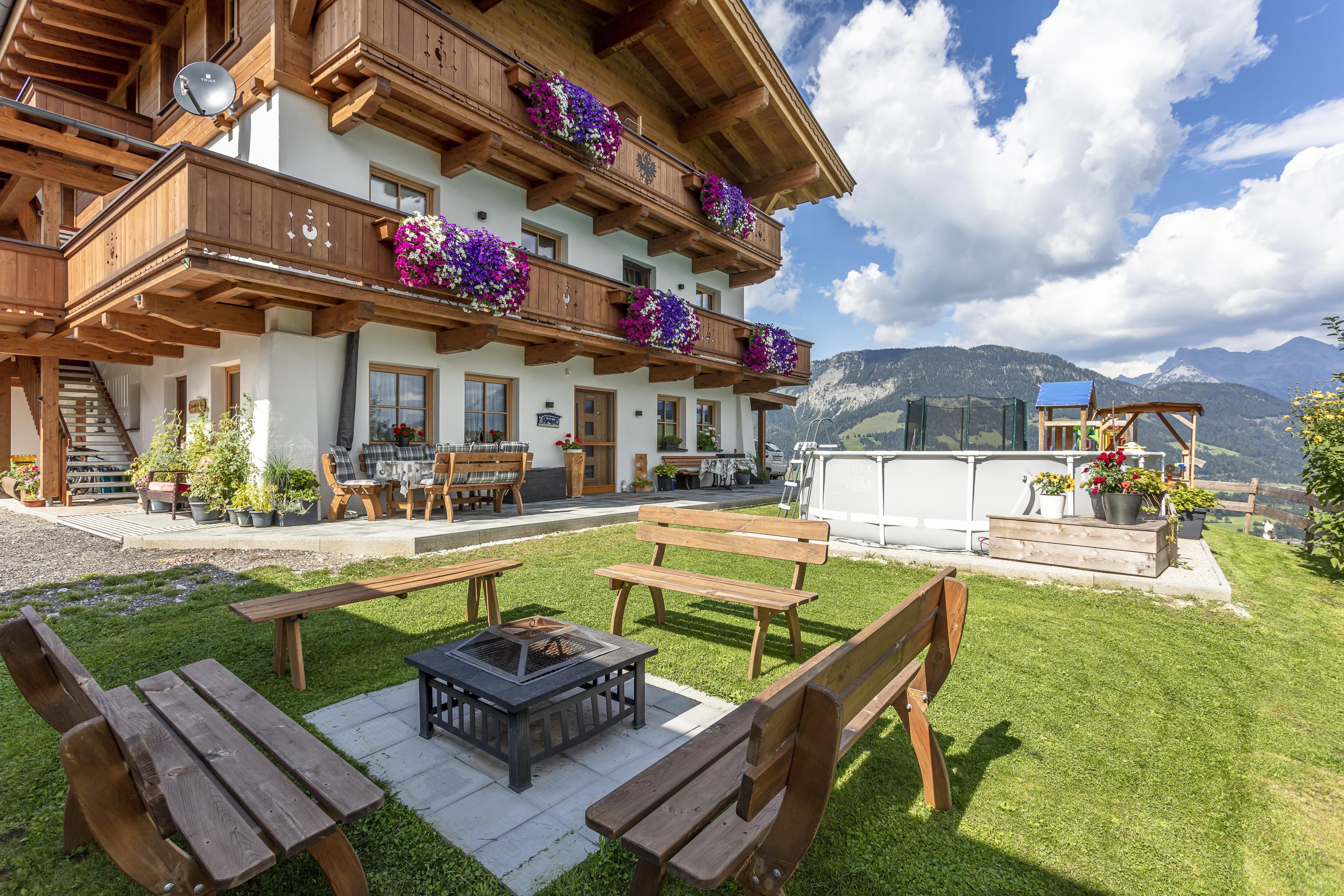 A cozy wooden building with balconies adorned with flowers. In the foreground, there is a fire pit and benches on a green meadow.
