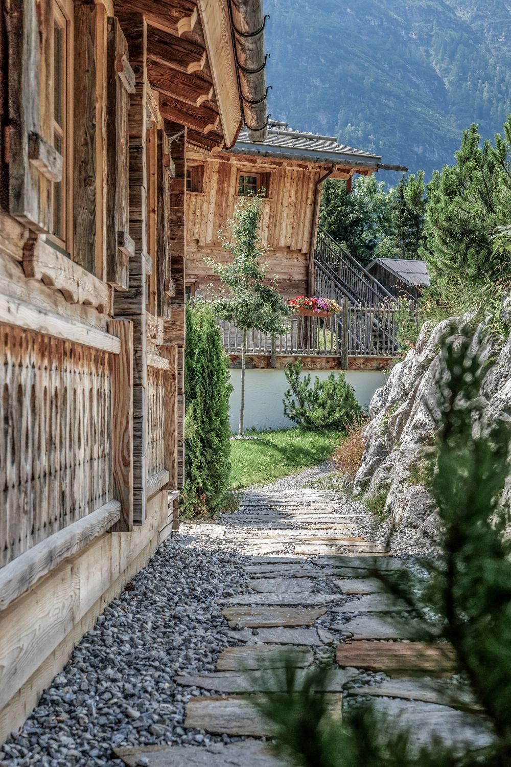 A picturesque path made of stones leads past chalets. Surrounded by green plants and mountains, the scene conveys a tranquil atmosphere.
