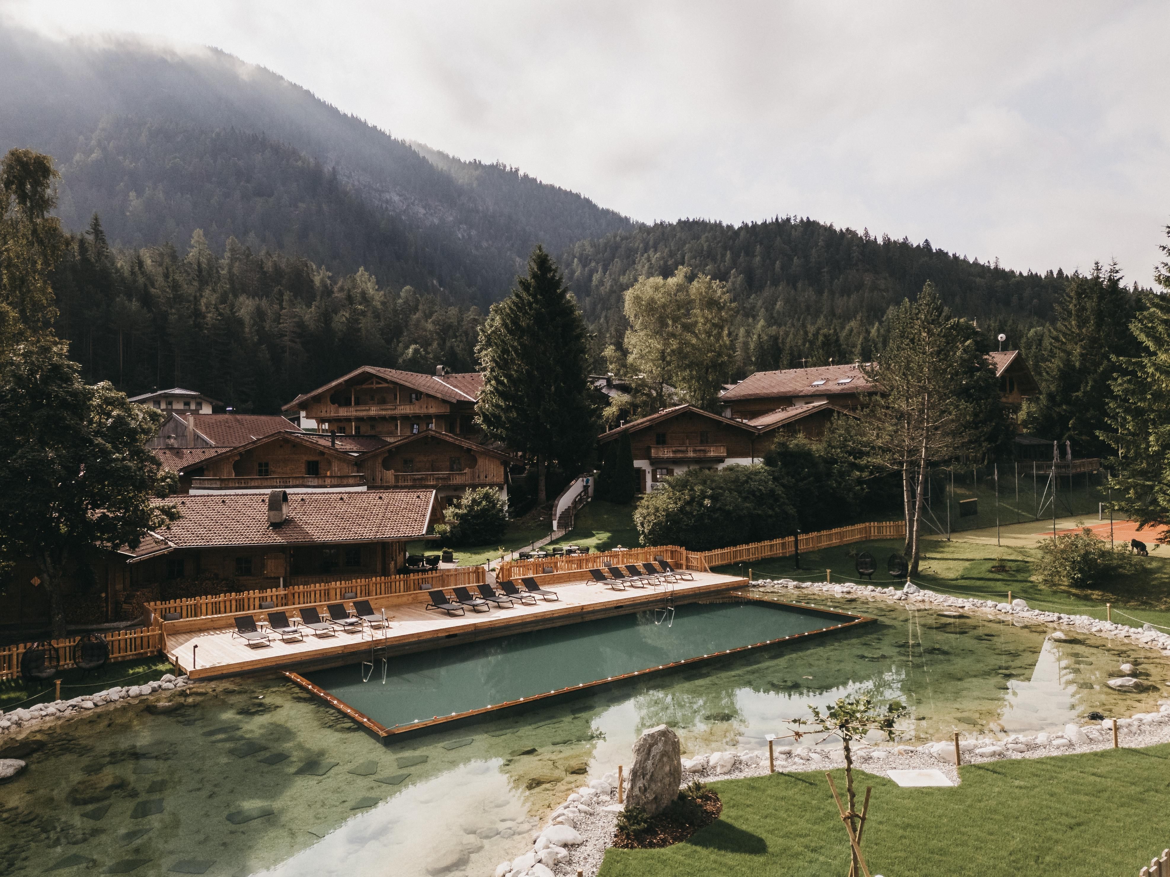 A picturesque mountain landscape with a small swimming pond and cozy wooden chalets. The surroundings are surrounded by trees and mountains, creating a relaxed atmosphere.