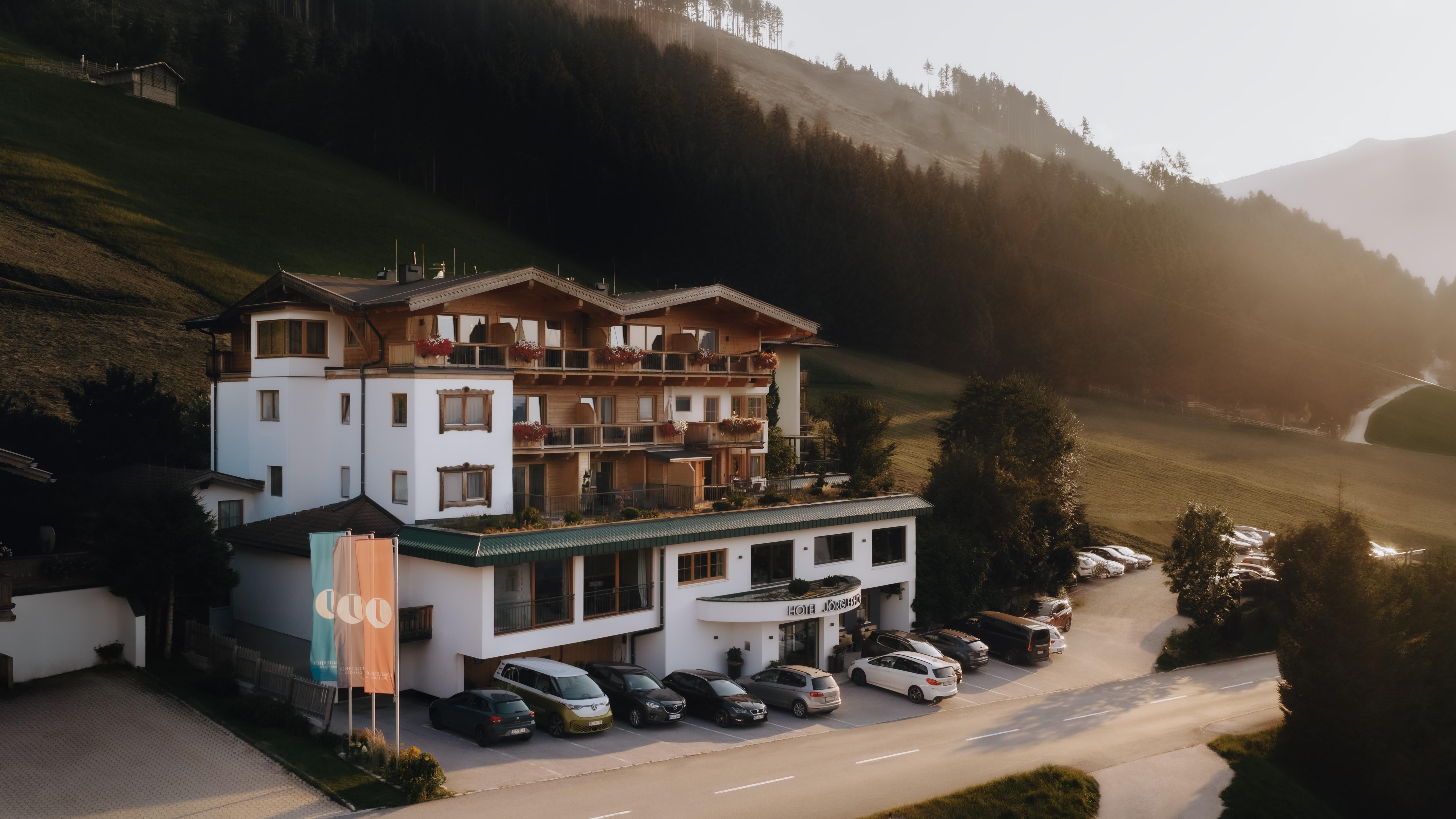 A cozy hotel in the mountains with several balconies and large windows. In the foreground, parked cars and a quiet street landscape.