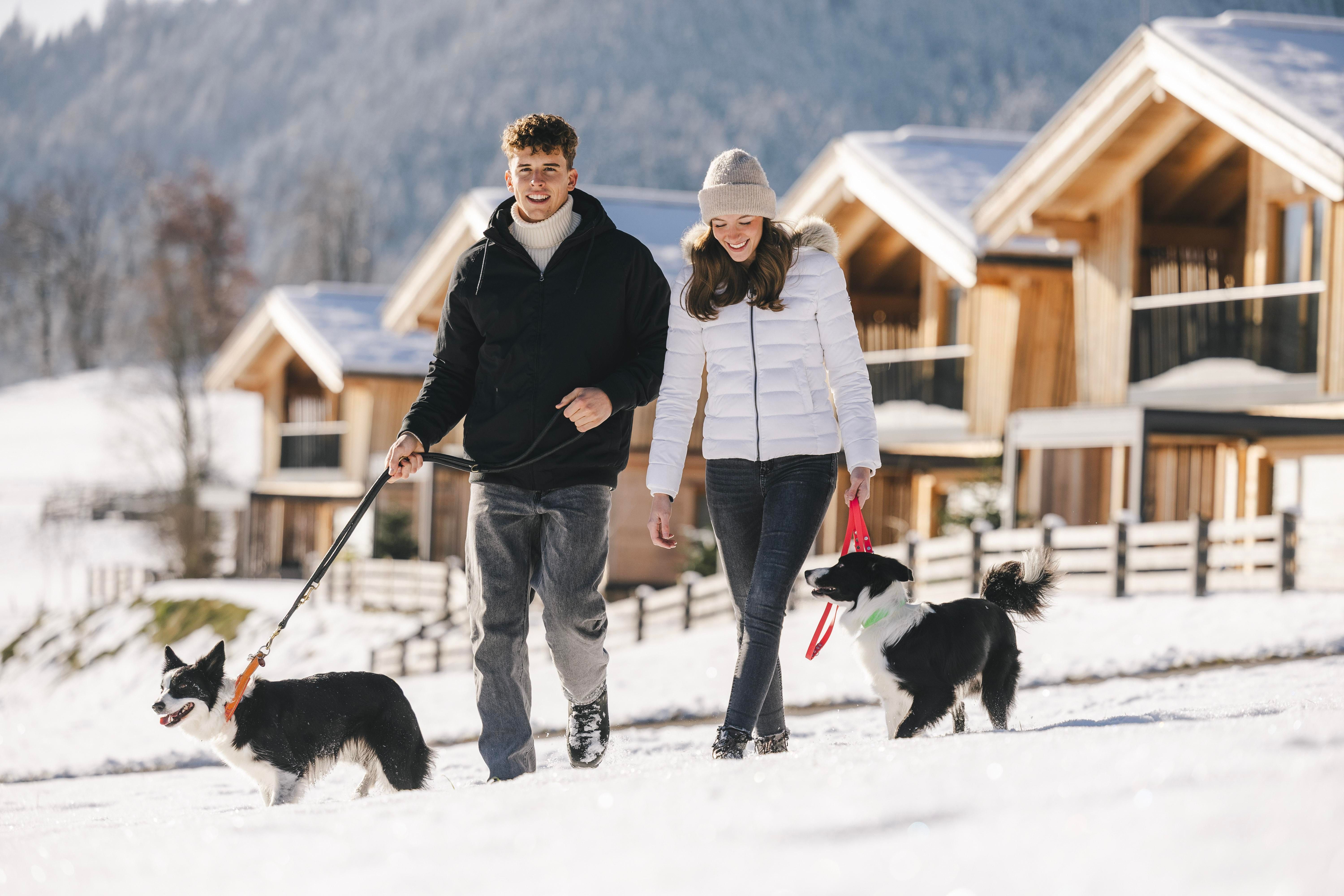 A couple is walking with two dogs through a snowy area. In the background, modern wooden houses and mountains can be seen.