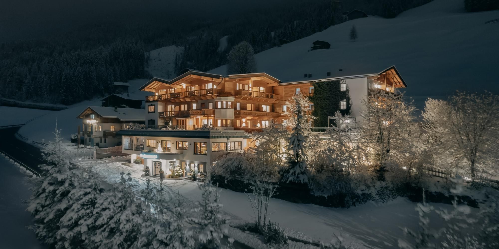 A charming hotel in a snowy landscape at night, brightly illuminated by warm lights. In the background, snow-covered mountains and trees are visible.