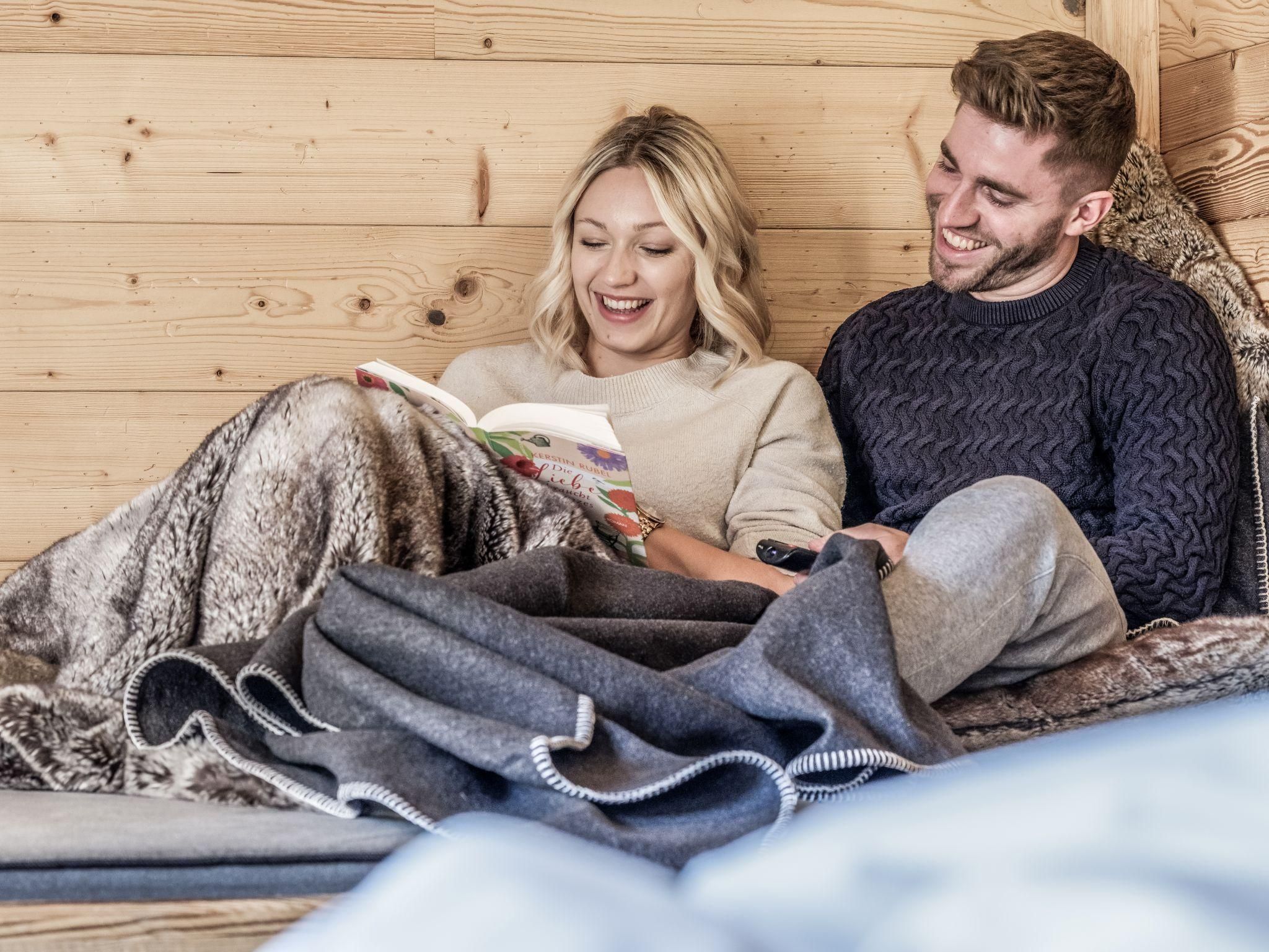 A couple sits comfortably in a chalet and reads a book together. They are covered with a blanket and smile relaxed.