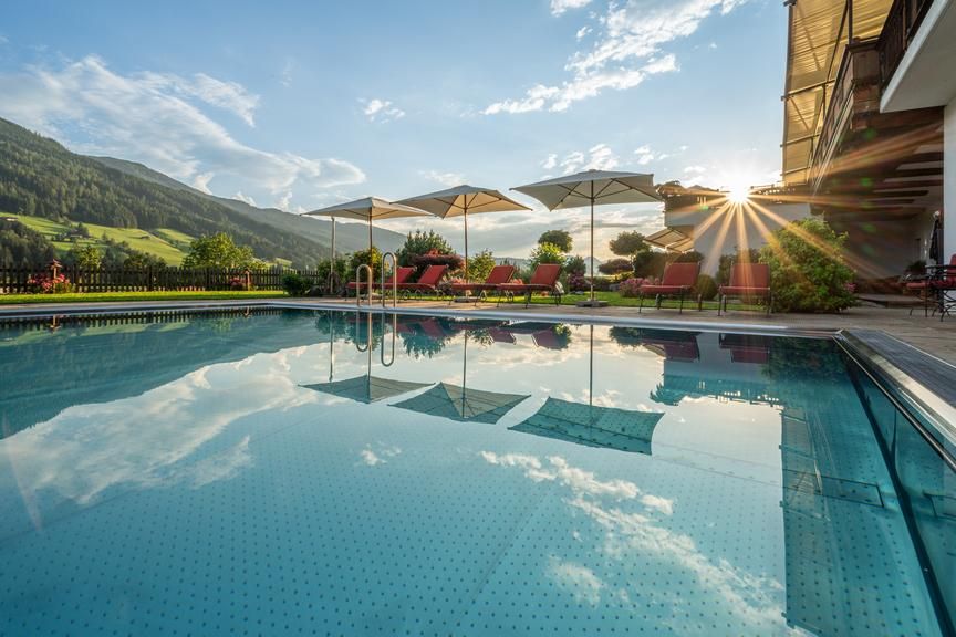 A beautifully designed pool in the Alps, surrounded by green hills. Sun rays shine through the clouds and reflect on the water.