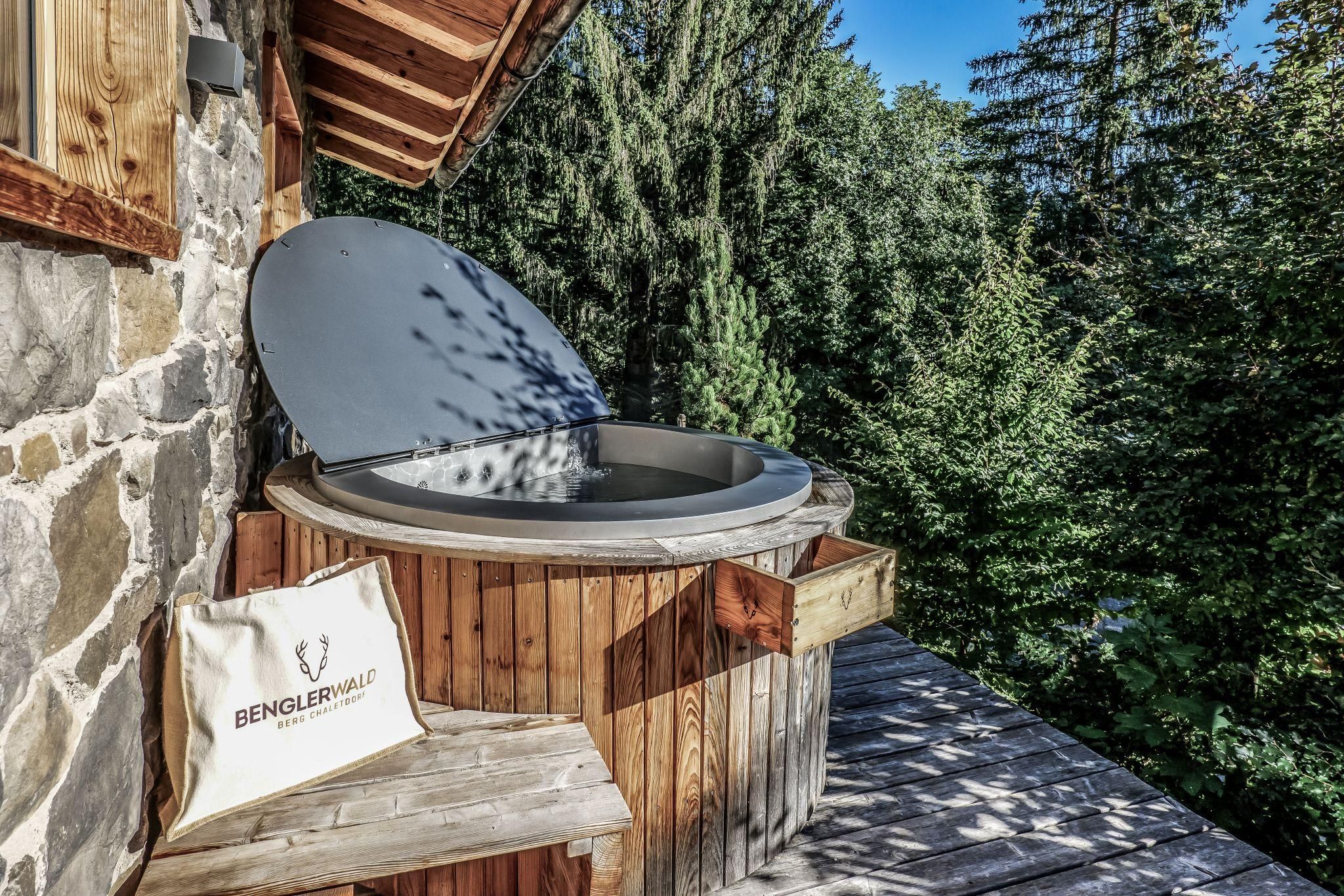 A relaxation area with a modern hot pot on a wooden terrace. Surrounded by green trees and blue sky, it offers a peaceful atmosphere.
