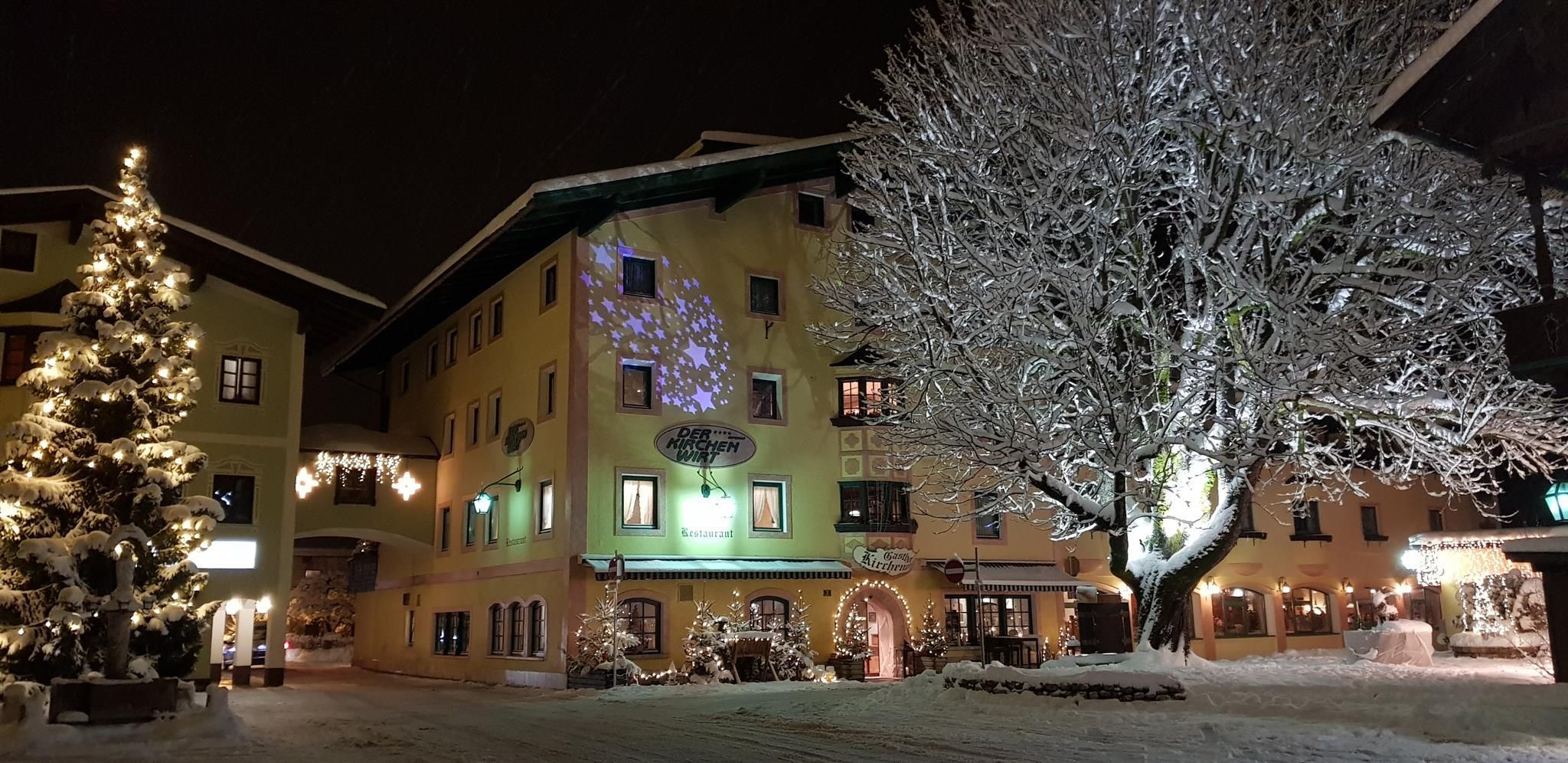 A cozy spot in the snow at night, illuminated by Christmas lights. A large Christmas tree stands on the left, while a decorated tree glows on the right.