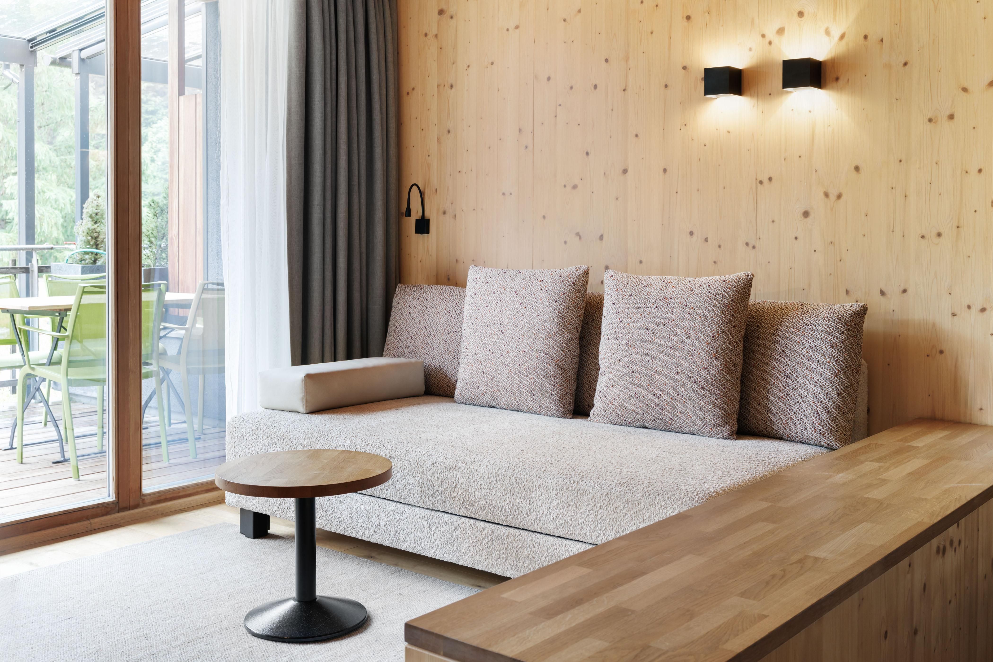 A modern living room with a beige sofa and large cushions. The room features wood paneling and large windows that overlook a balcony with chairs.