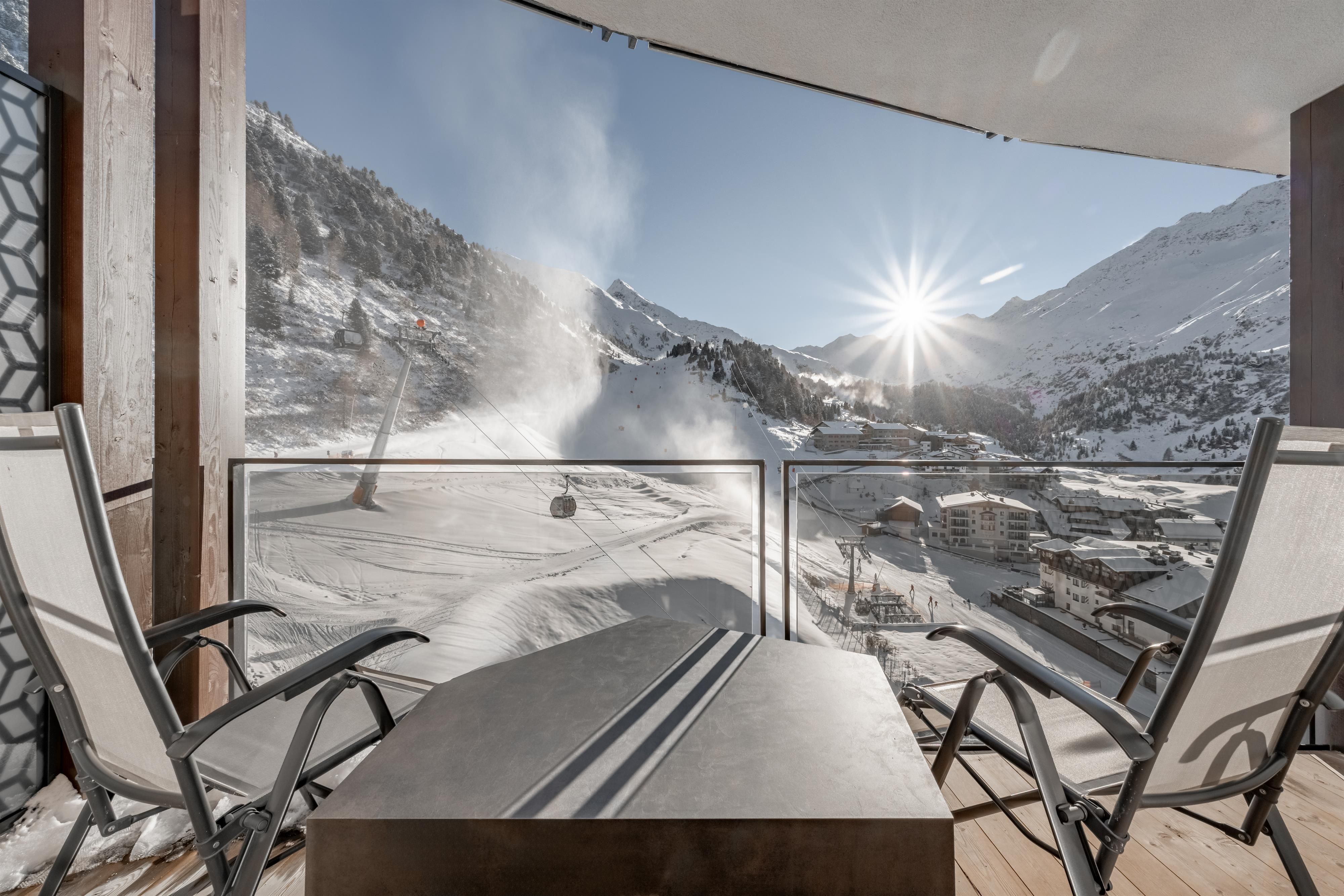 A terrace with chairs offers a view of a snowy mountain landscape. The sun is shining over the mountains and there is a ski lift station nearby.