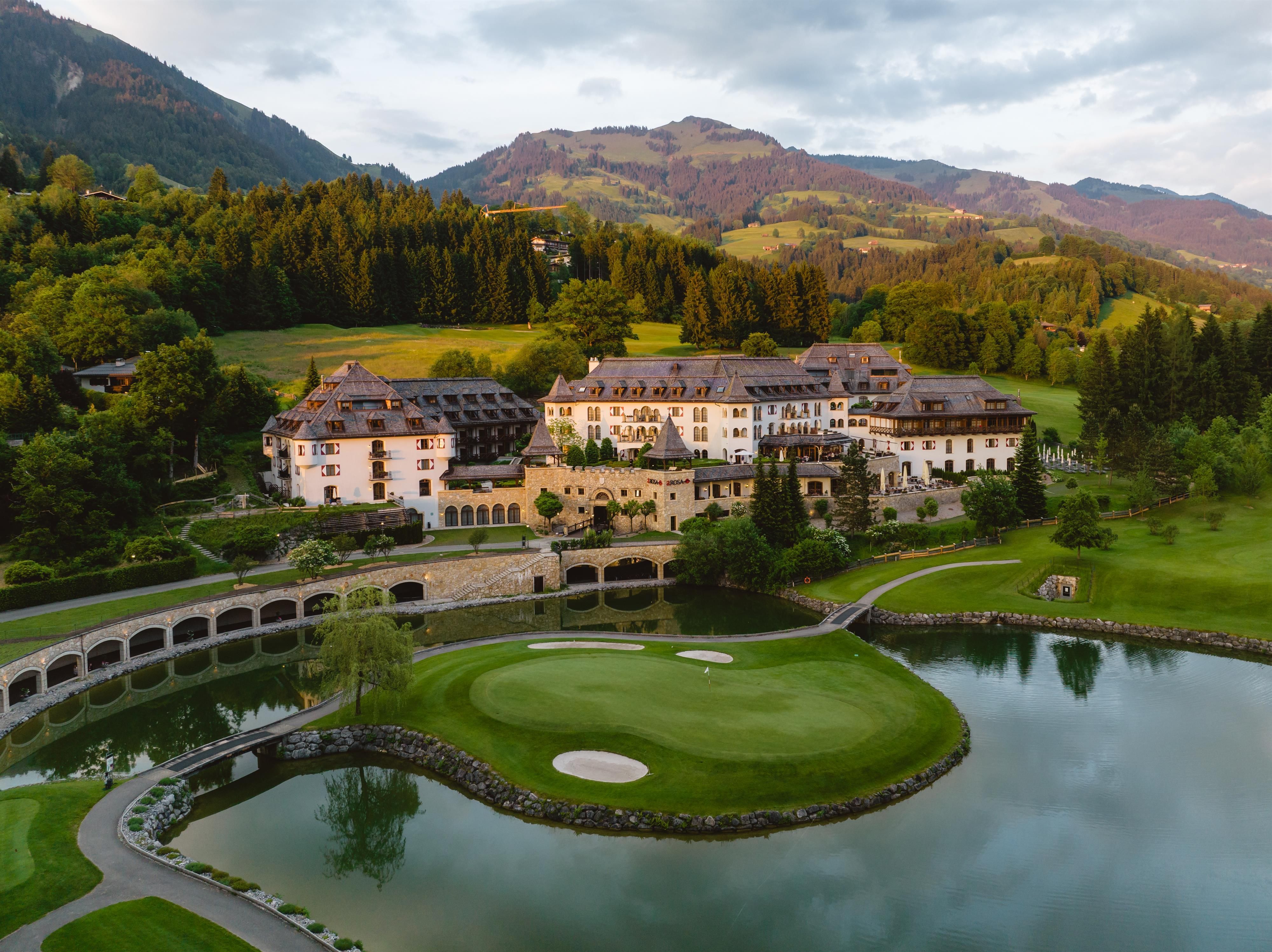 An elegant hotel amidst a green landscape and mountainous backdrop. A golf course and a tranquil lake surround the estate.