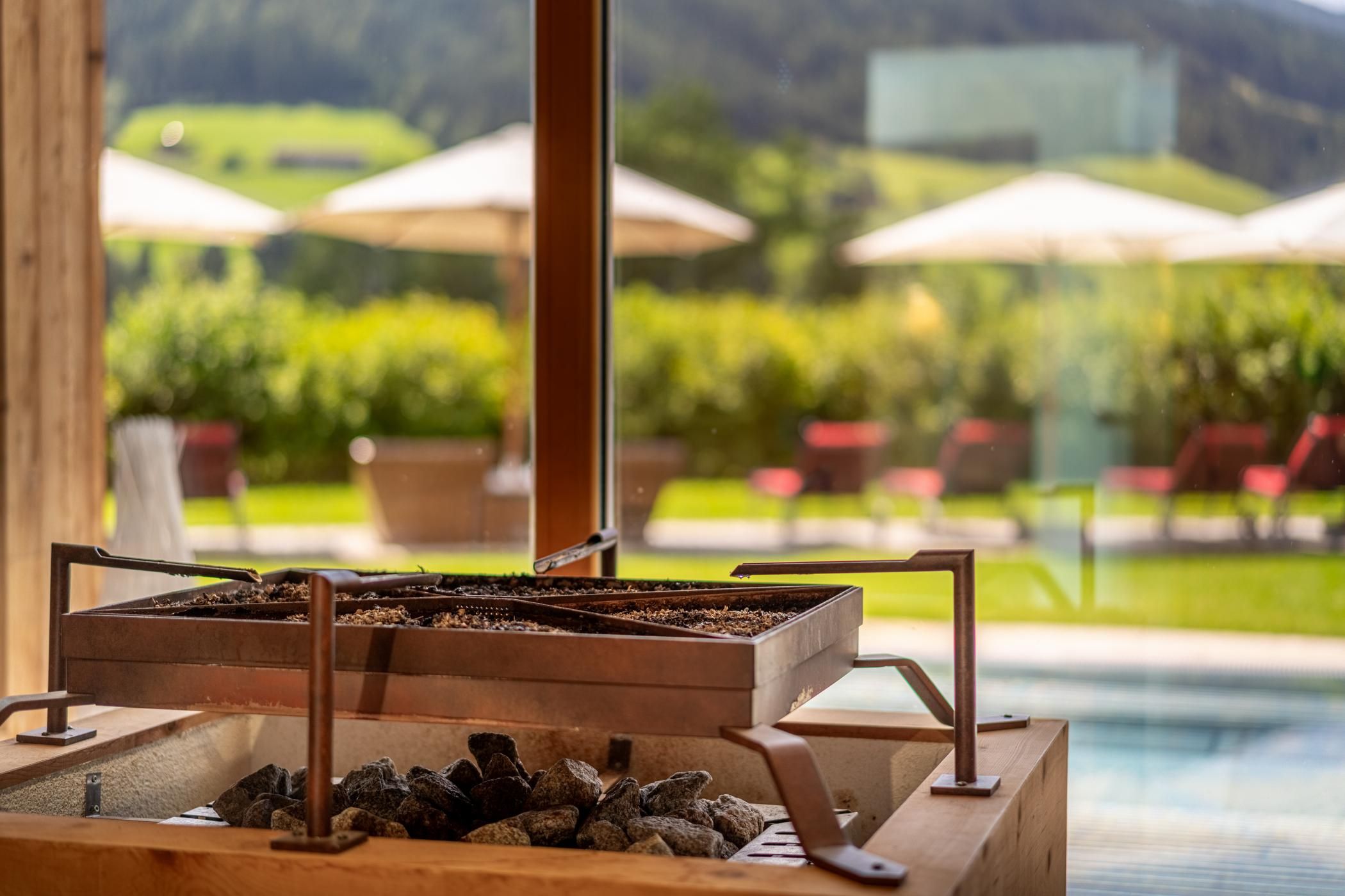 A modern sauna with a wooden and stone sauna stove. In the background, a green landscape and loungers with sun umbrellas can be seen.