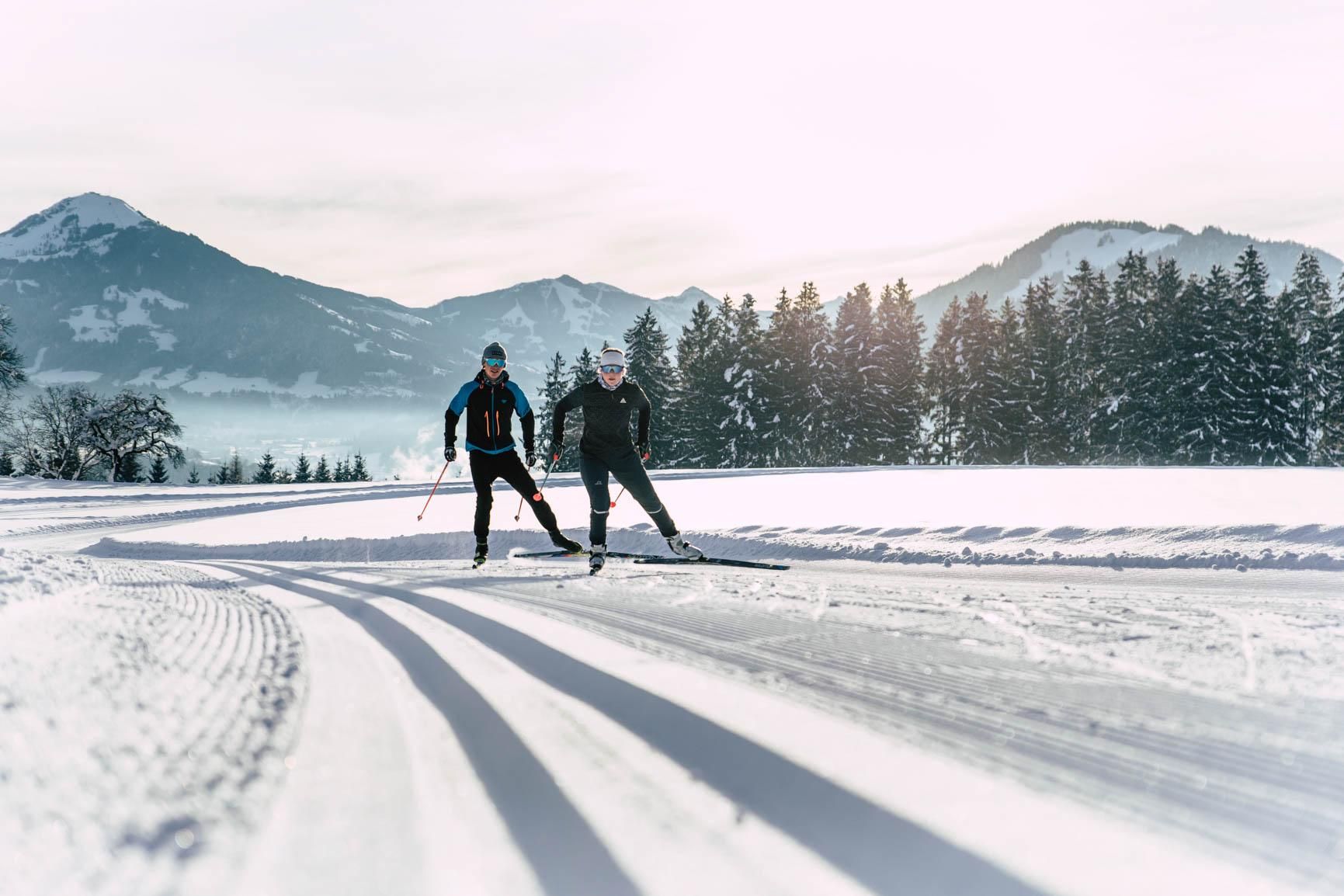 Two skiers glide on a snow-covered slope. In the background, mountains and coniferous trees can be seen.