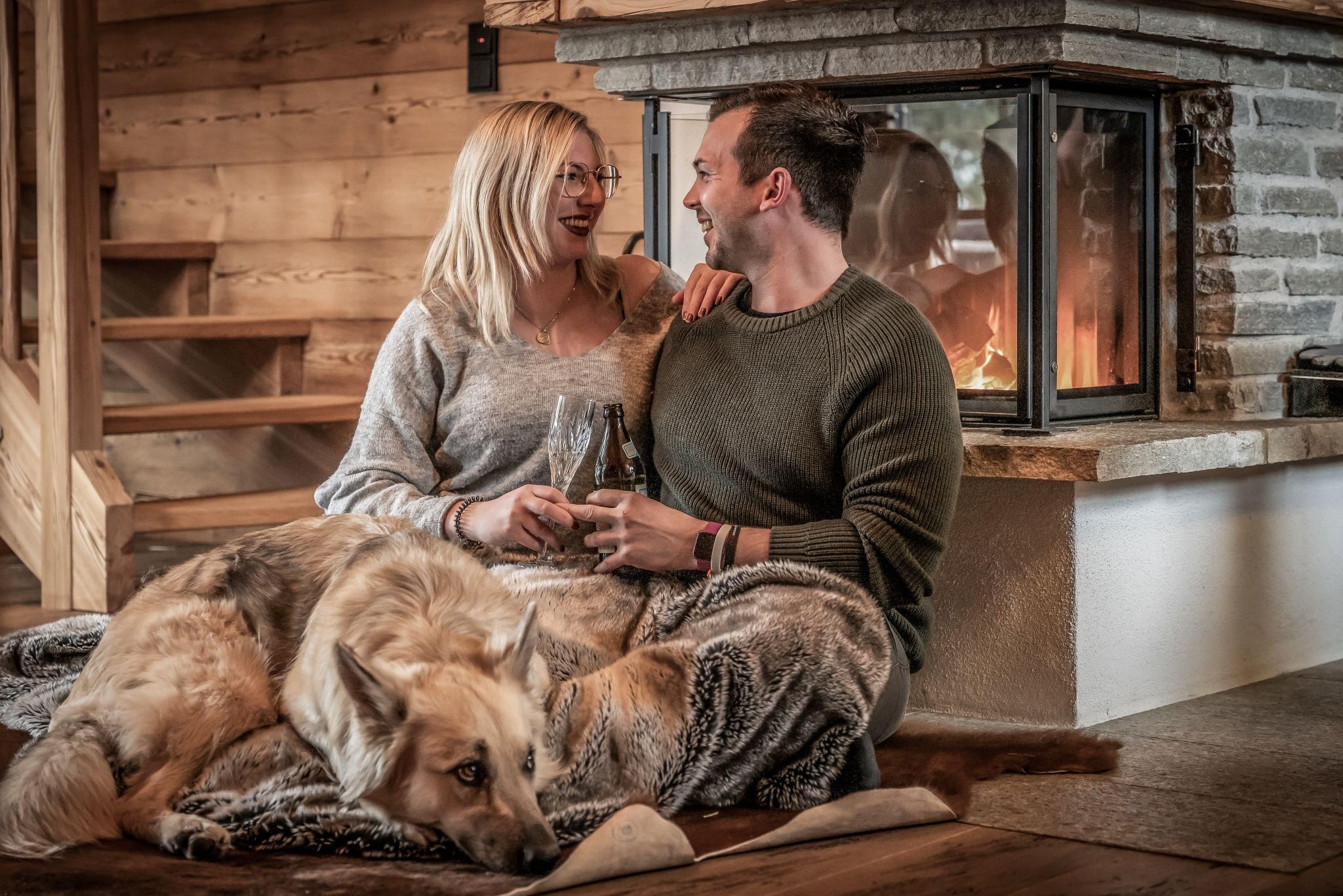 A couple sits comfortably in front of a fireplace and enjoys a relaxed moment. A dog lies relaxed next to them on a blanket.