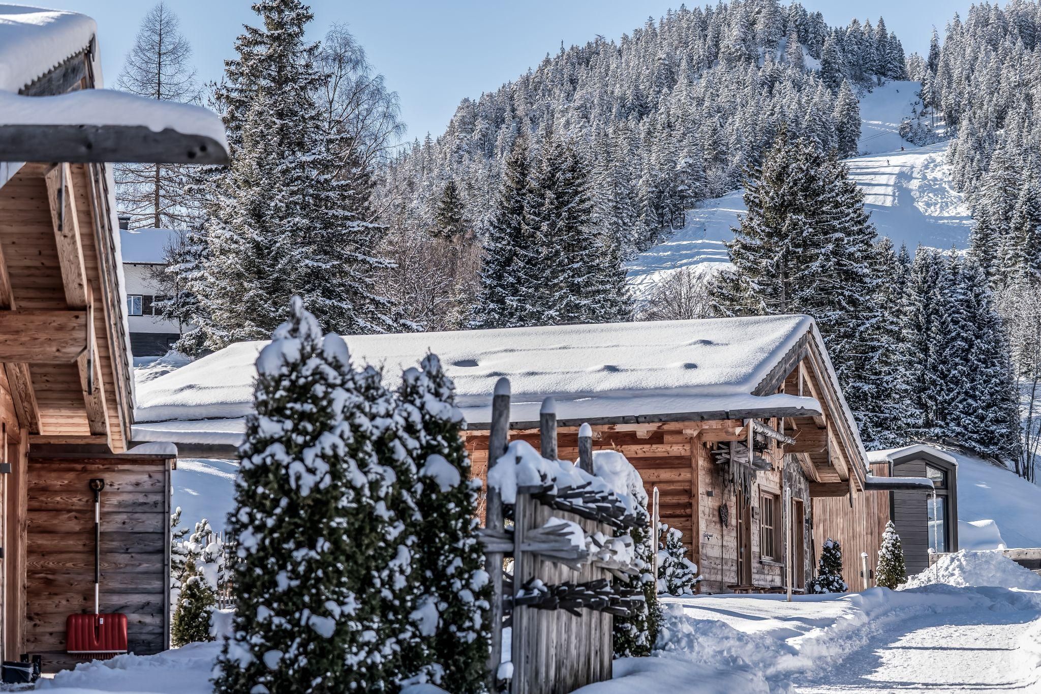A picturesque winter village with snow-covered chalets and tall fir trees. In the background, one can see gentle, snowy hills.