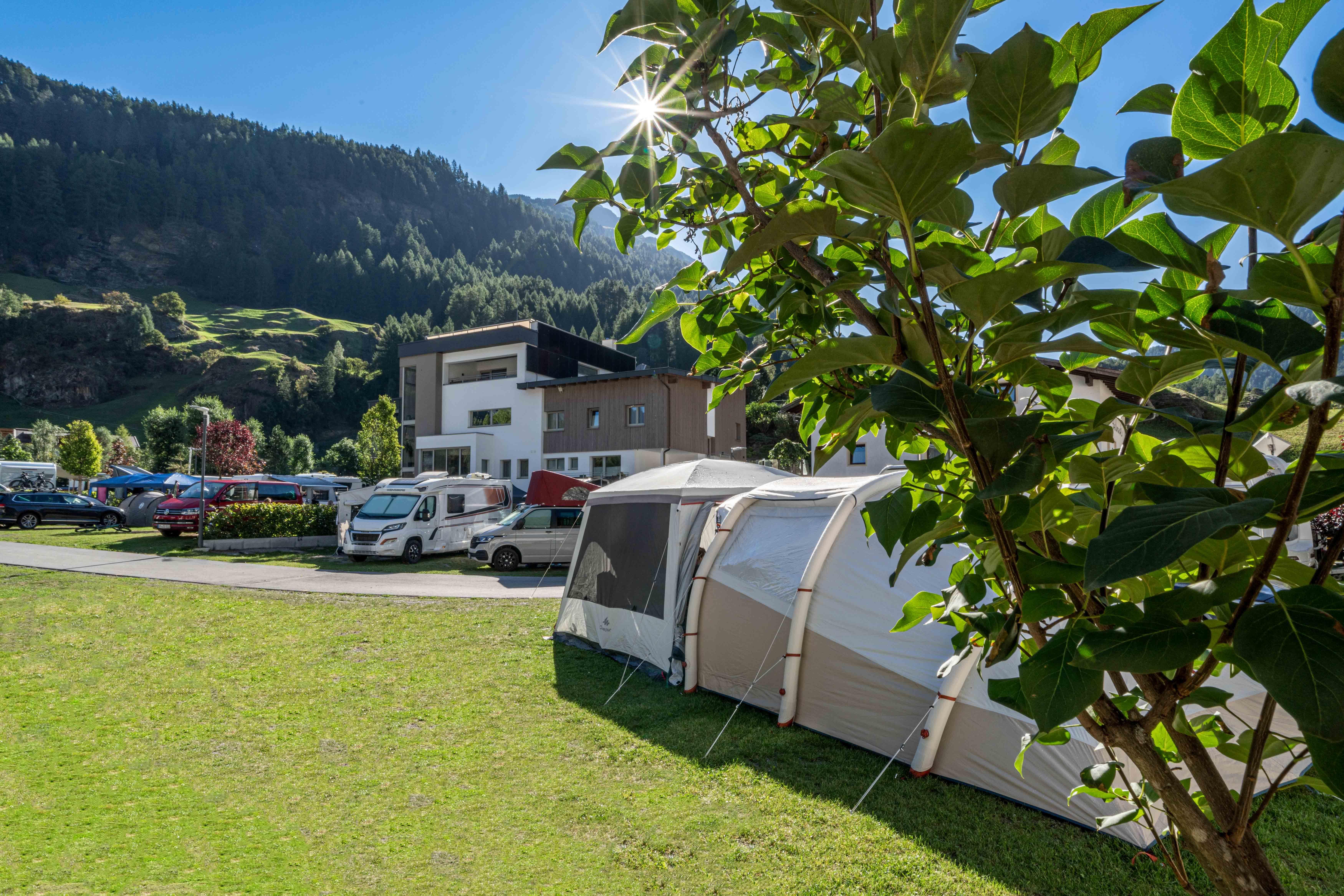 A cozy campsite with tent houses and motorhomes. In the background, green hills and a modern building can be seen.