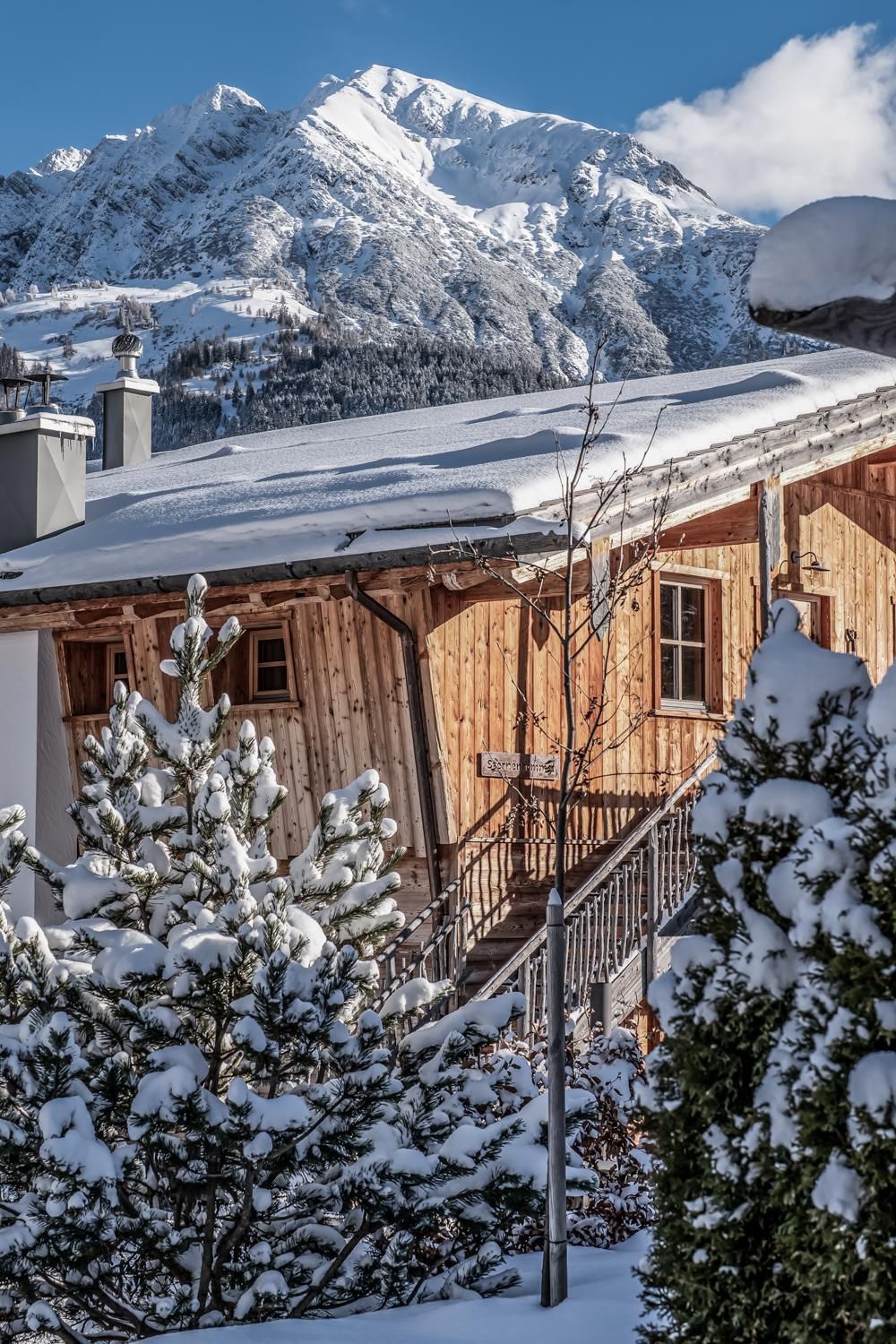 A cozy chalet in the snow, surrounded by snow-covered trees. In the background, majestic mountains are visible under a clear blue sky.