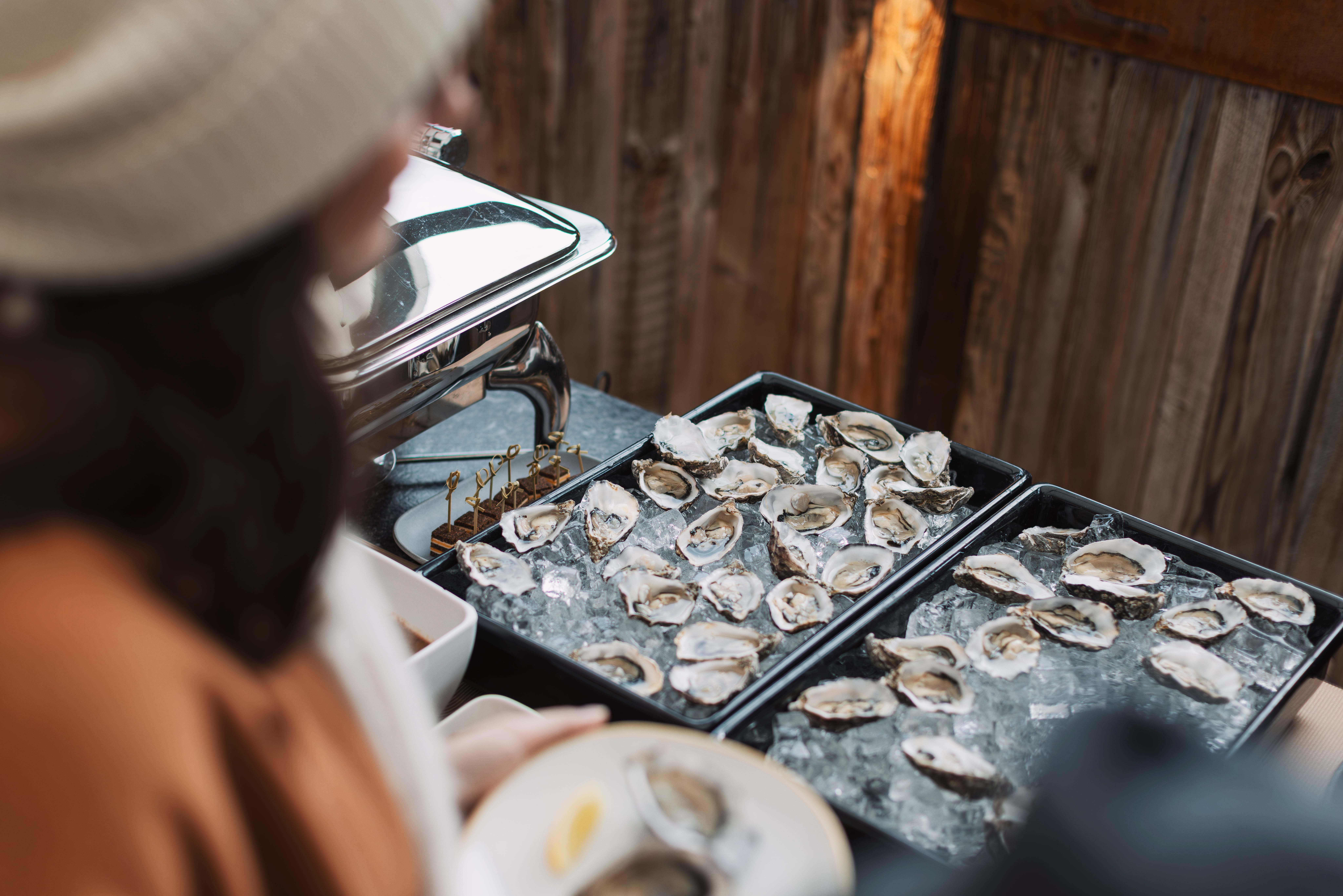 A selection of fresh oysters on ice, served in several bowls. In the foreground, a person can be seen holding a plate with a lemon.