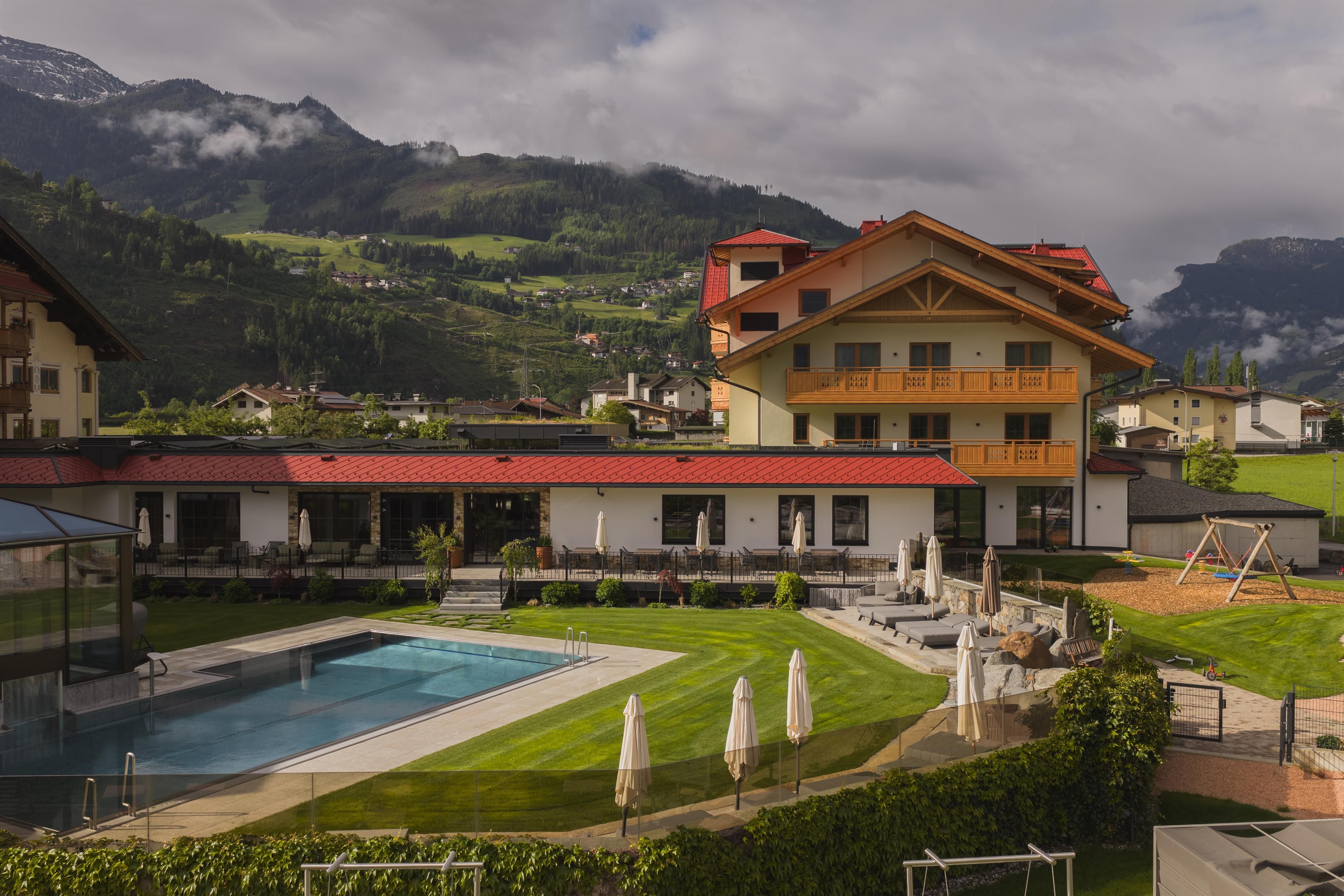 A hotel with beautiful architecture and an outdoor pool. The surroundings are green and surrounded by mountains.