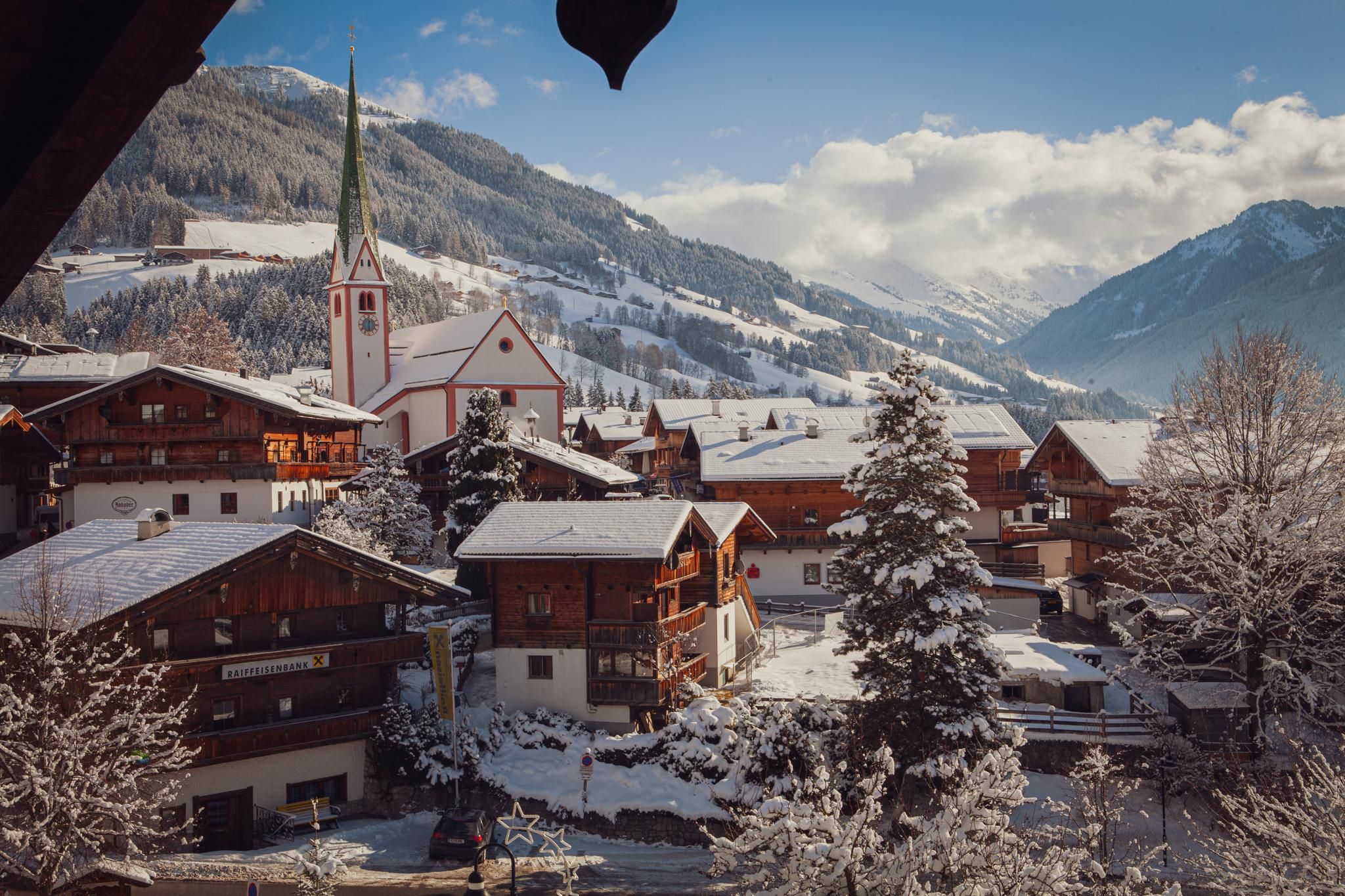 A picturesque village in winter with snow-covered houses and a church. The surrounding mountains are visible in the background, under a clear sky.