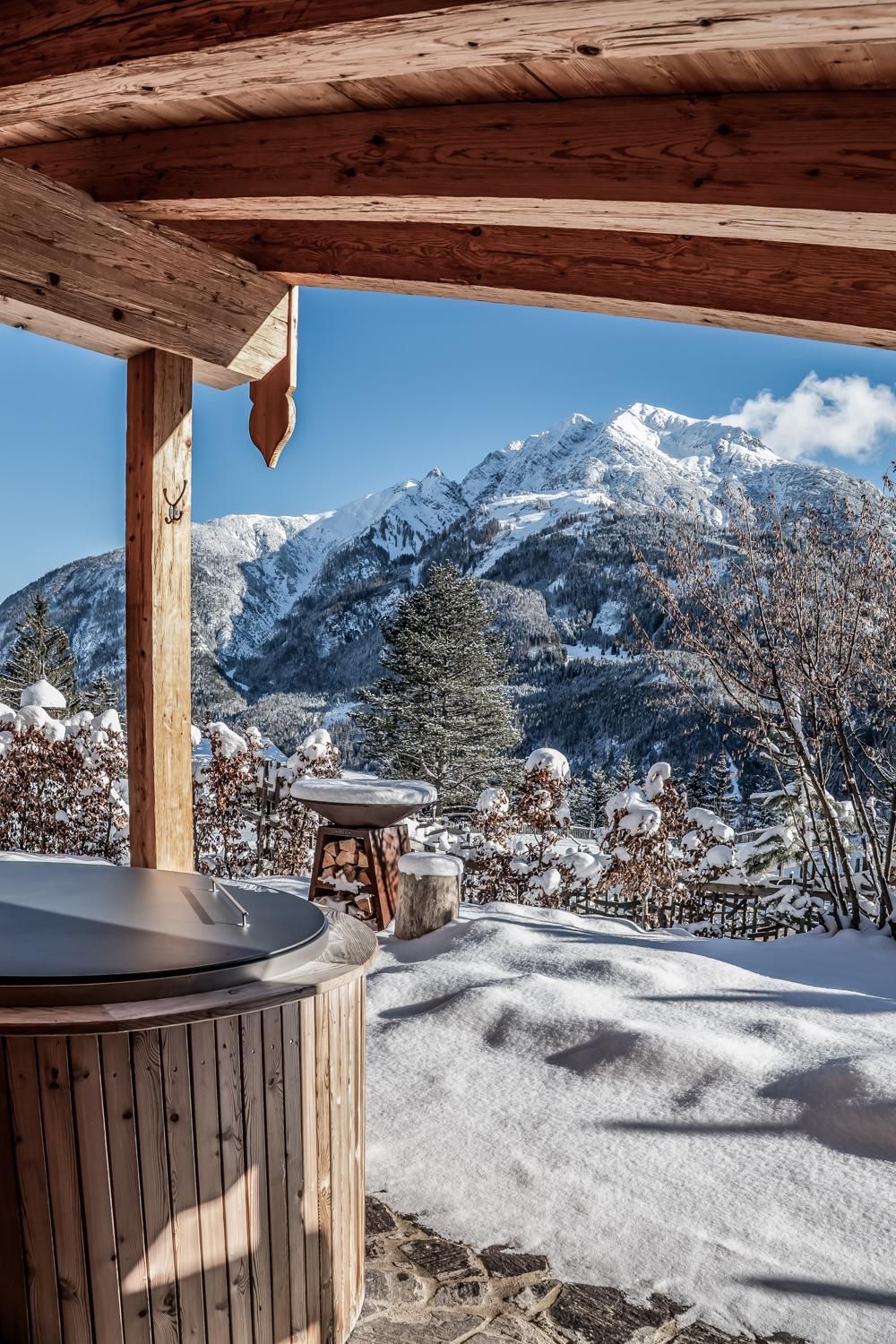 A cozy outdoor area with a view of snow-covered mountains. The ground is covered with fresh snow and the sun is shining in the clear sky.