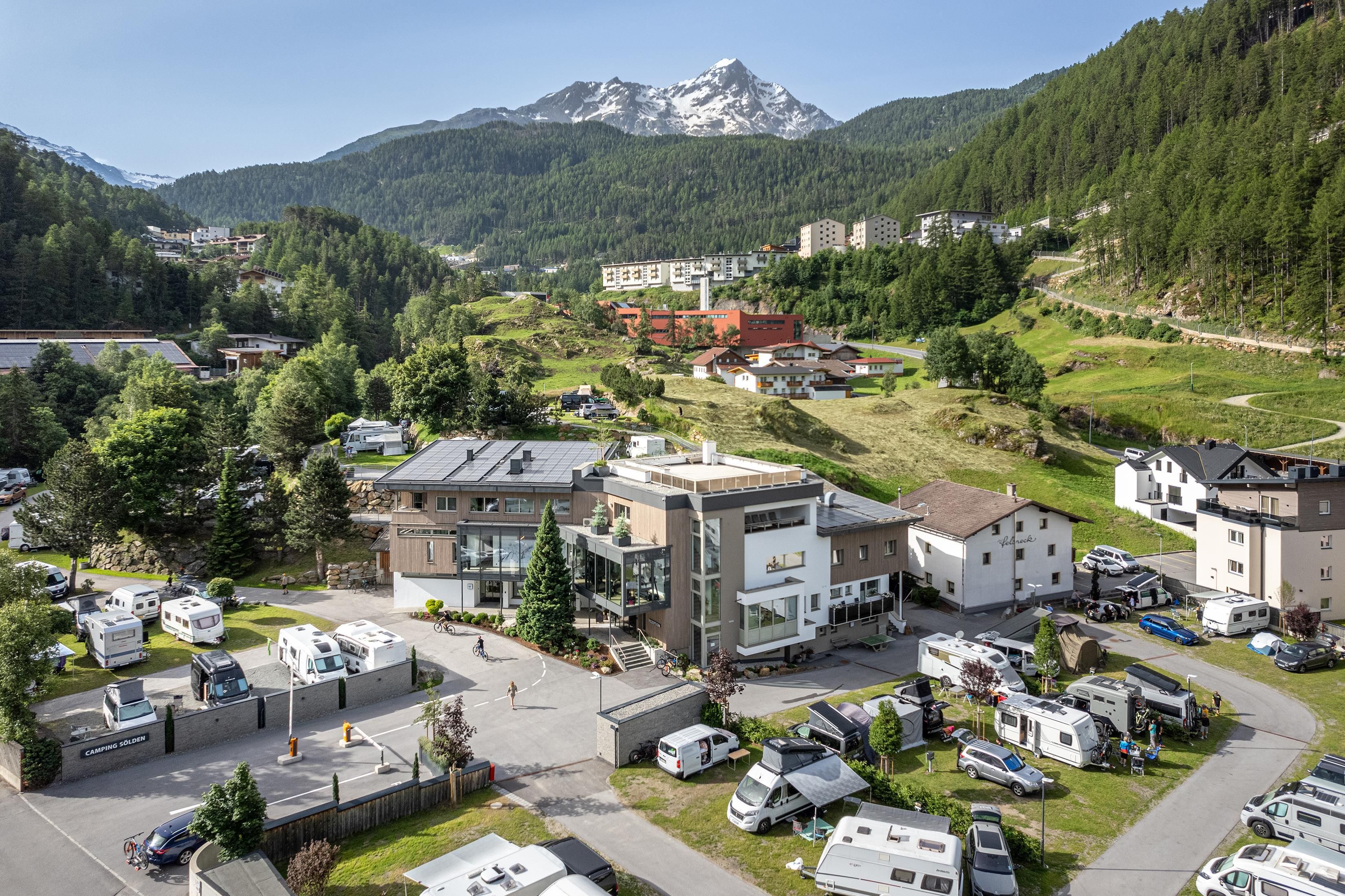 A picturesque mountain landscape with snow-capped peaks in the background. In the foreground, you can see a campsite with RVs and cozy buildings.