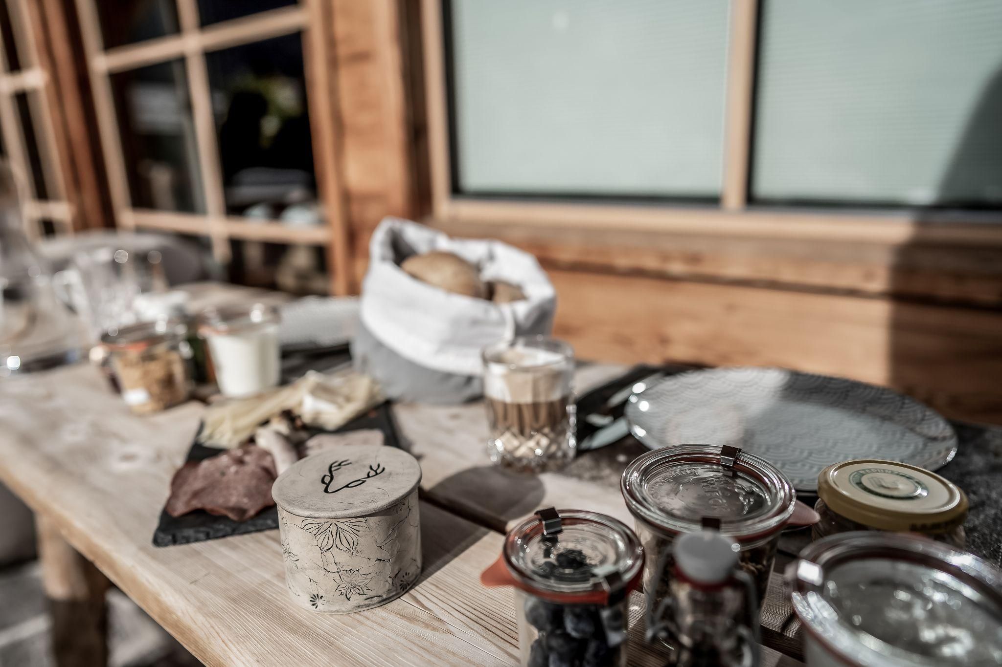 A rustic table with various breakfast foods, including bread, cheese, and jam. The scene is bright and inviting, surrounded by wood.