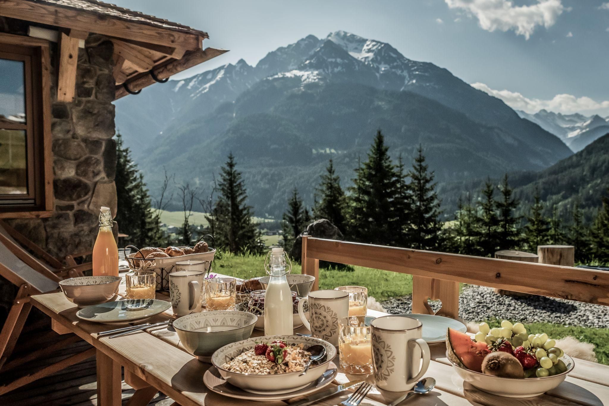 A set table with a lavish breakfast is on a terrace. In the background, majestic mountains and a clear sky can be seen.
