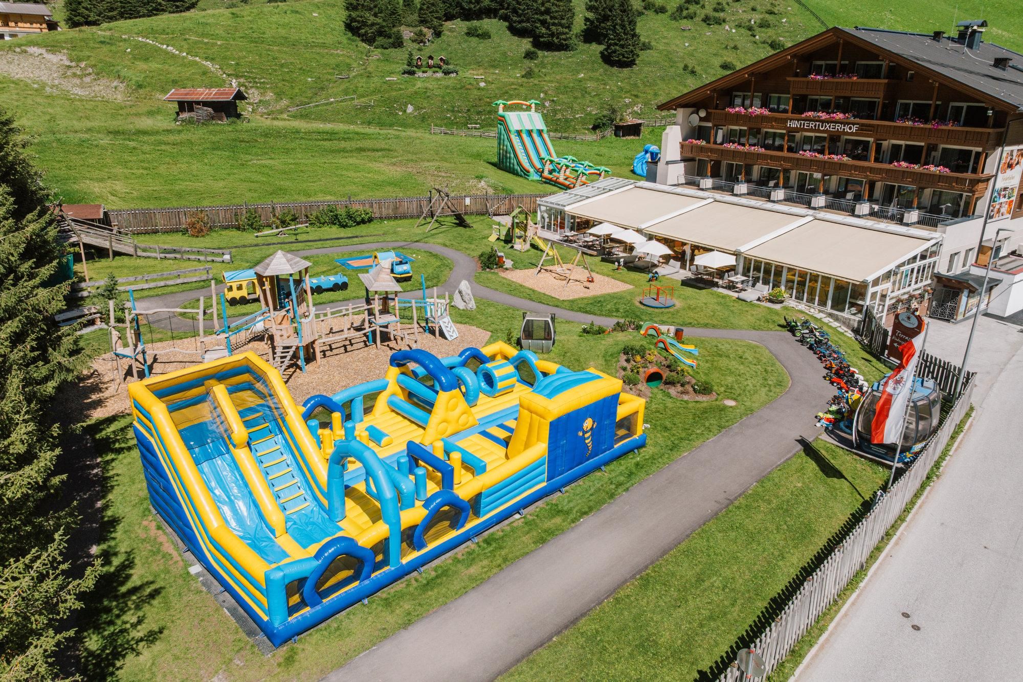 A large, colorful bouncy castle playground with slides and various play equipment. In the background, there is a hotel with a terrace and green spaces.