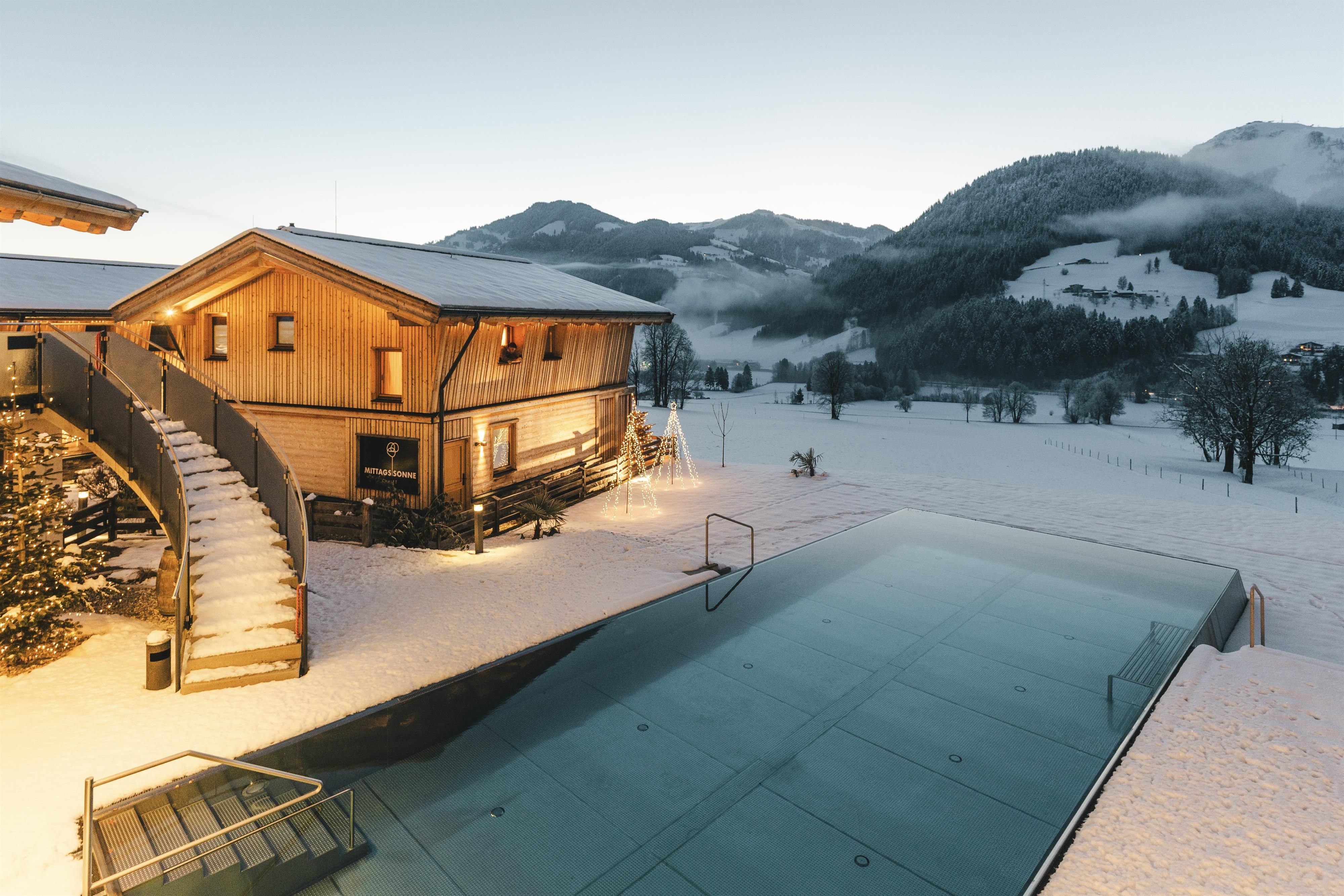 A modern wooden house in the snow with a large window front. In the foreground, there is an almost empty swimming pool, and in the background, there are gentle, snow-covered hills.