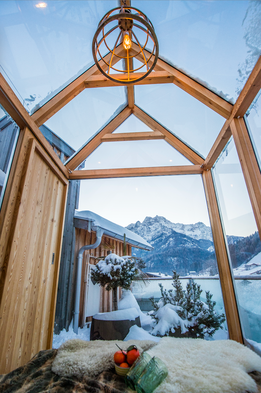 A cozy winter room with a glass structure and wooden details. In the background, snow-covered mountains can be seen.