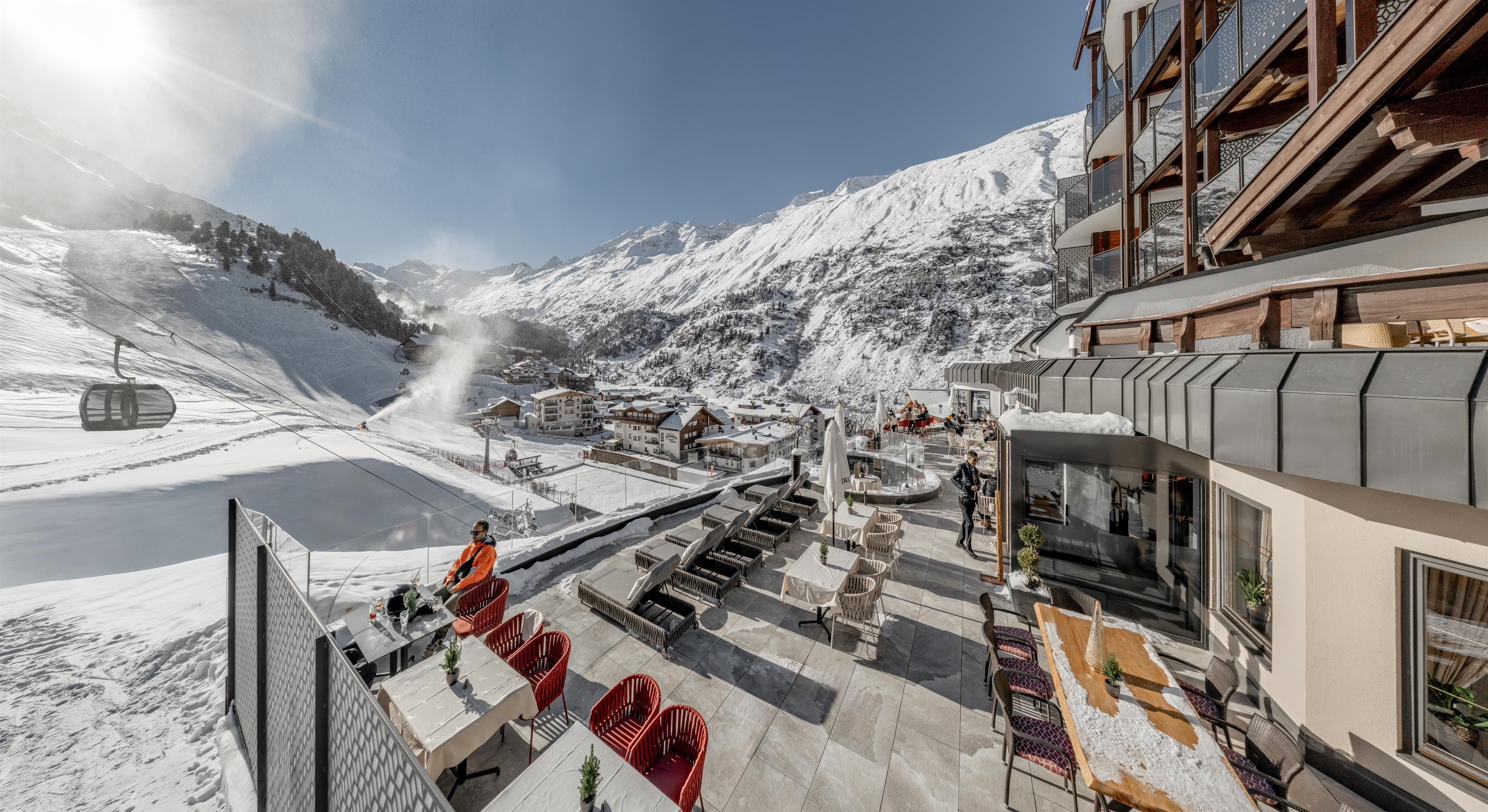 A beautiful view of an alpine landscape with snow-capped mountains. In the foreground, you can see cozy seating areas and a cable car that opens up the mountainous world.
