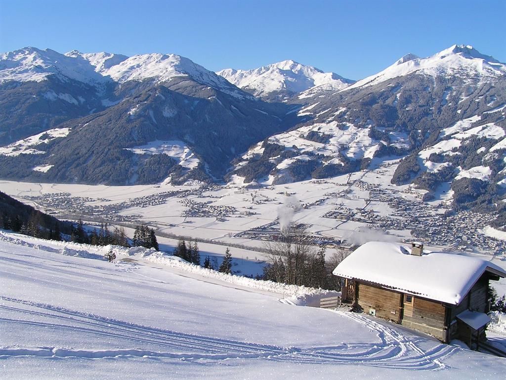 A beautiful winter landscape with snow-covered mountains and a small wooden house in the foreground. The sky is clear and blue, while the view into the valley, which is covered in snow, is captivating.