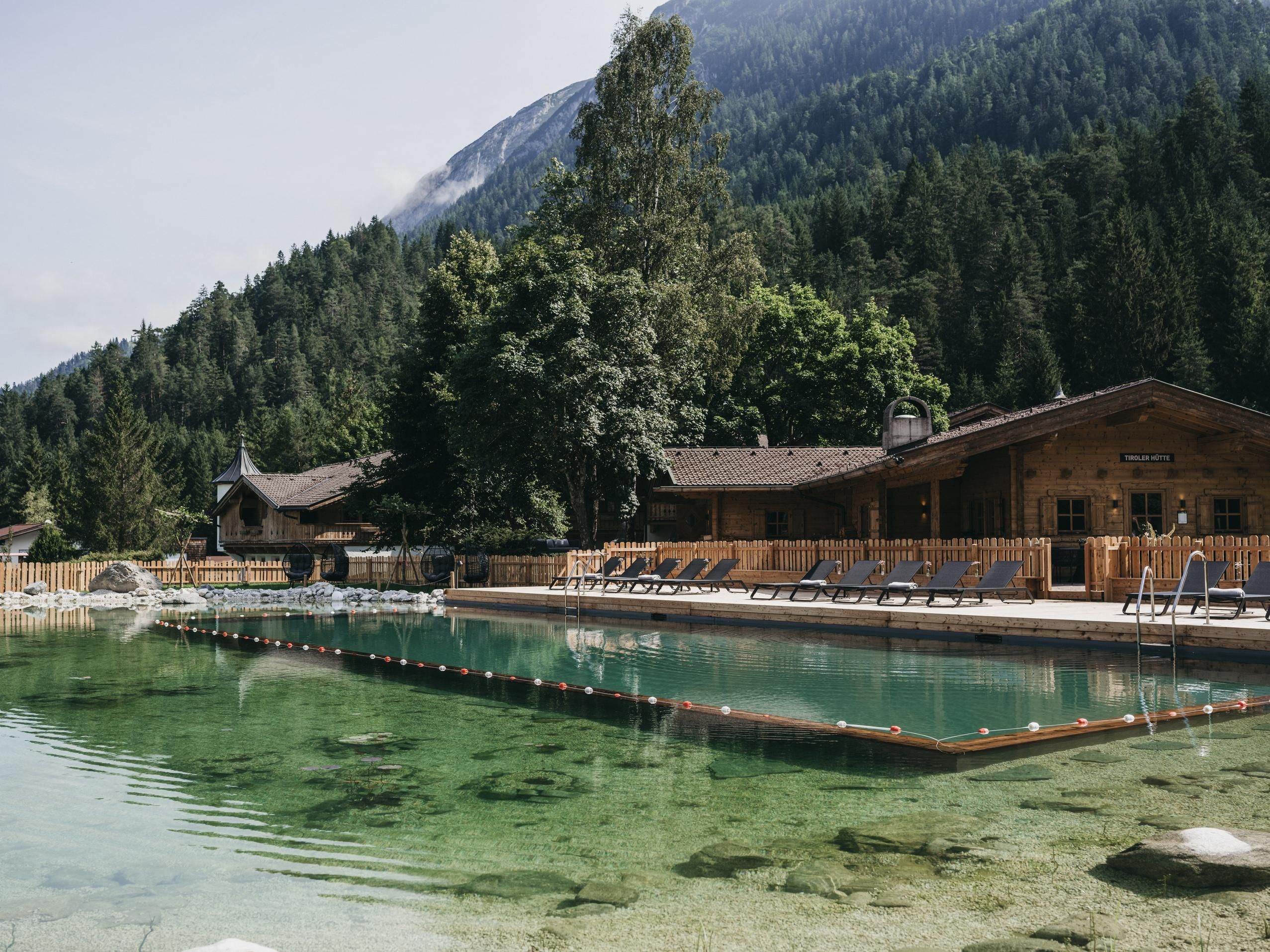 A quiet outdoor pool with clear water, surrounded by mountains and trees. In the background, wooden buildings are visible, creating a cozy atmosphere.