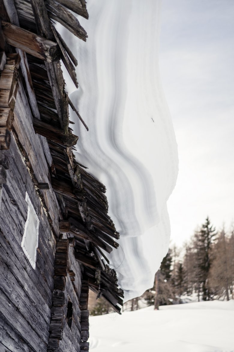 Snow covers the roof of a hut like a thick layer of icing on top of a cake.
