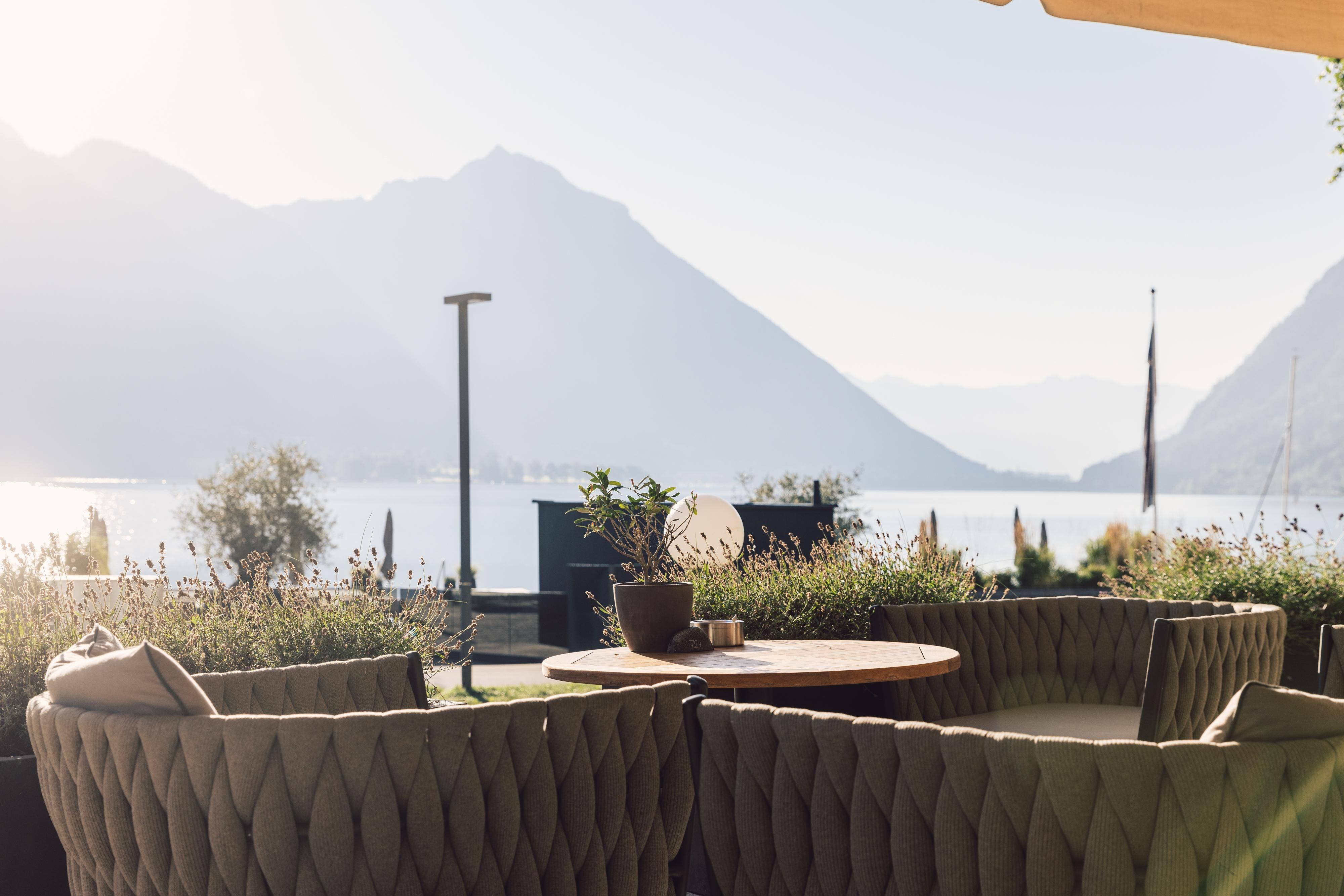A stylish terrace with cozy seating overlooks a tranquil lake. In the background, gentle mountains rise under a clear sky.