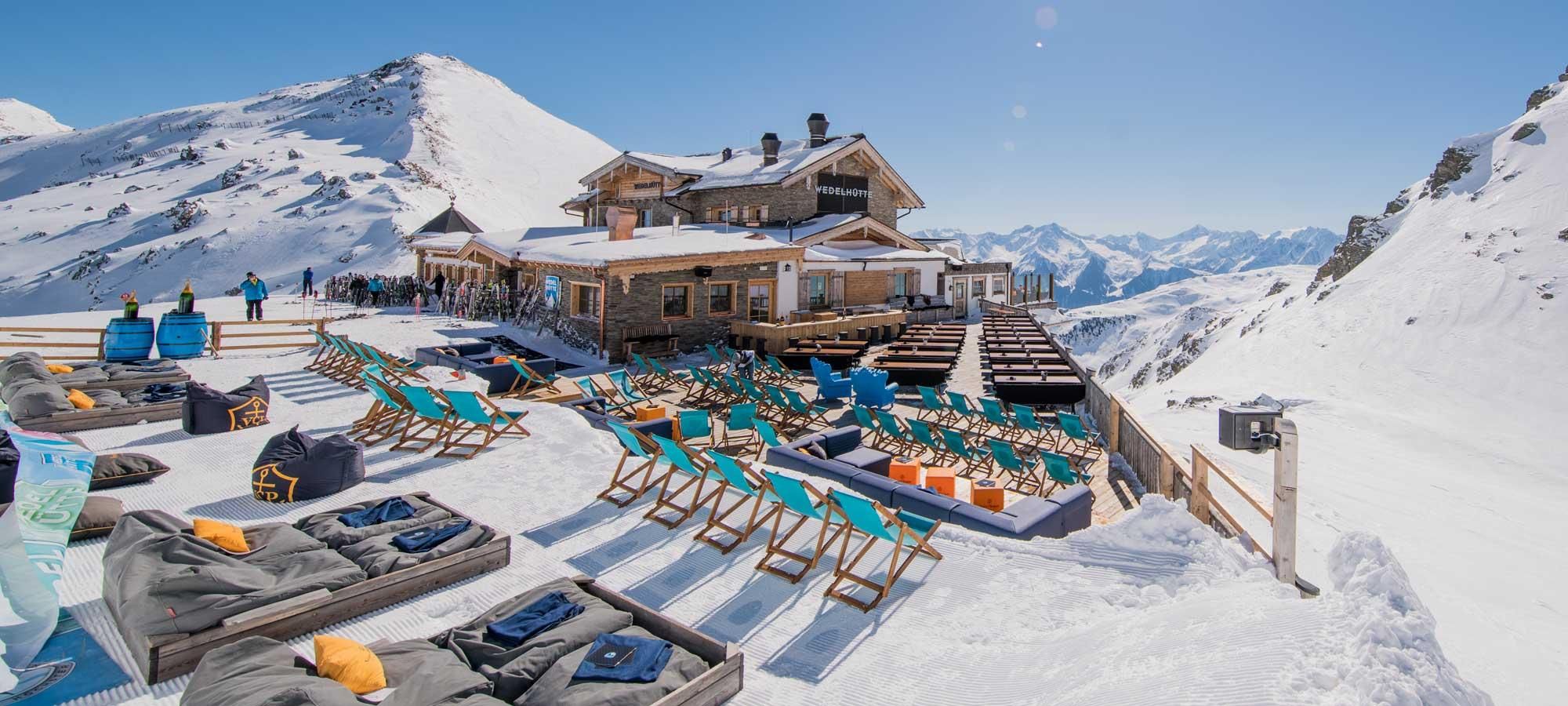 A picturesque mountain hut in the snow with cozy loungers and colorful seating. The surrounding mountains are clear and sunny.