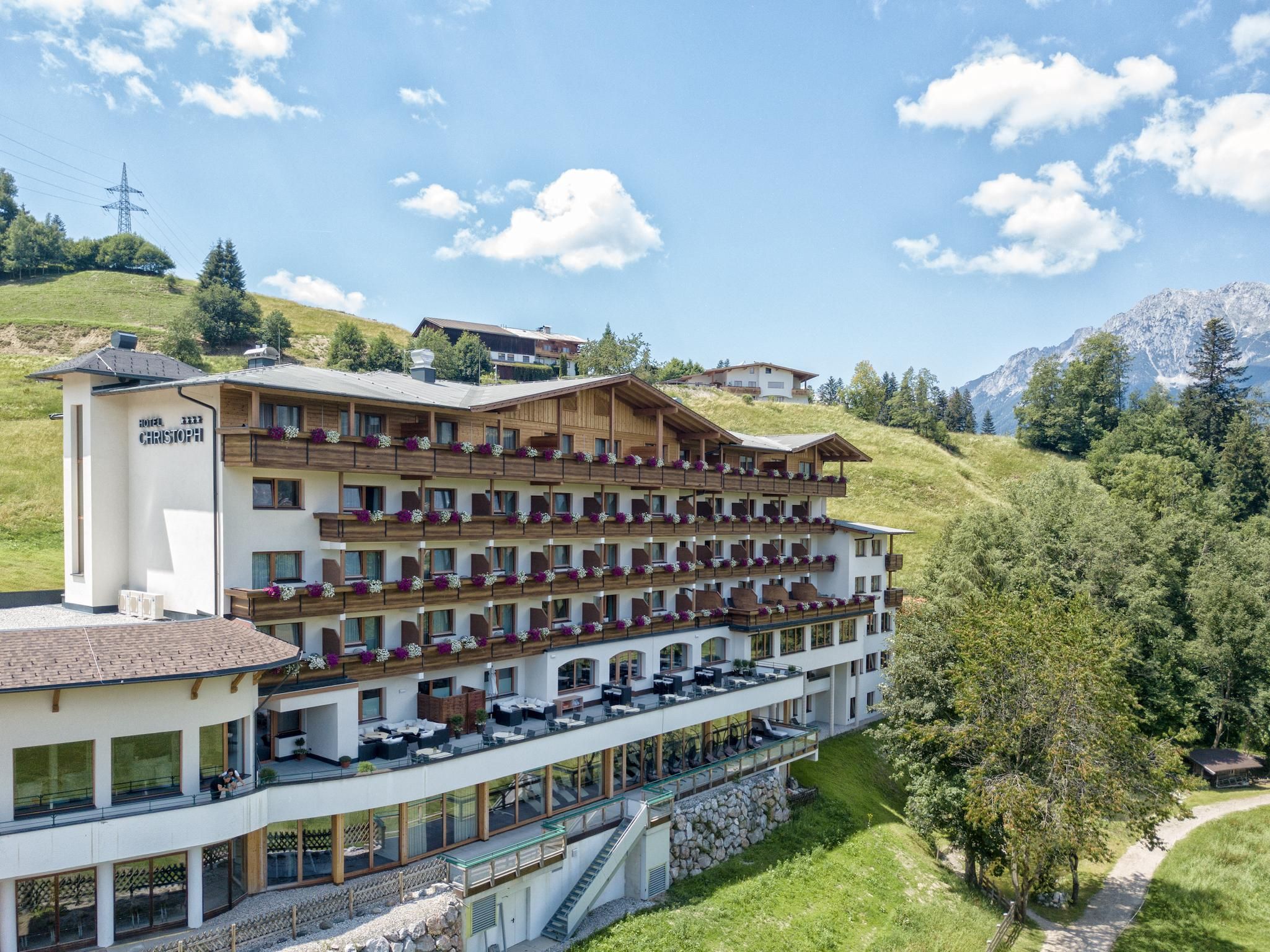 A modern hotel with several balconies, surrounded by green hills and mountains. The sky is clear and sunny.