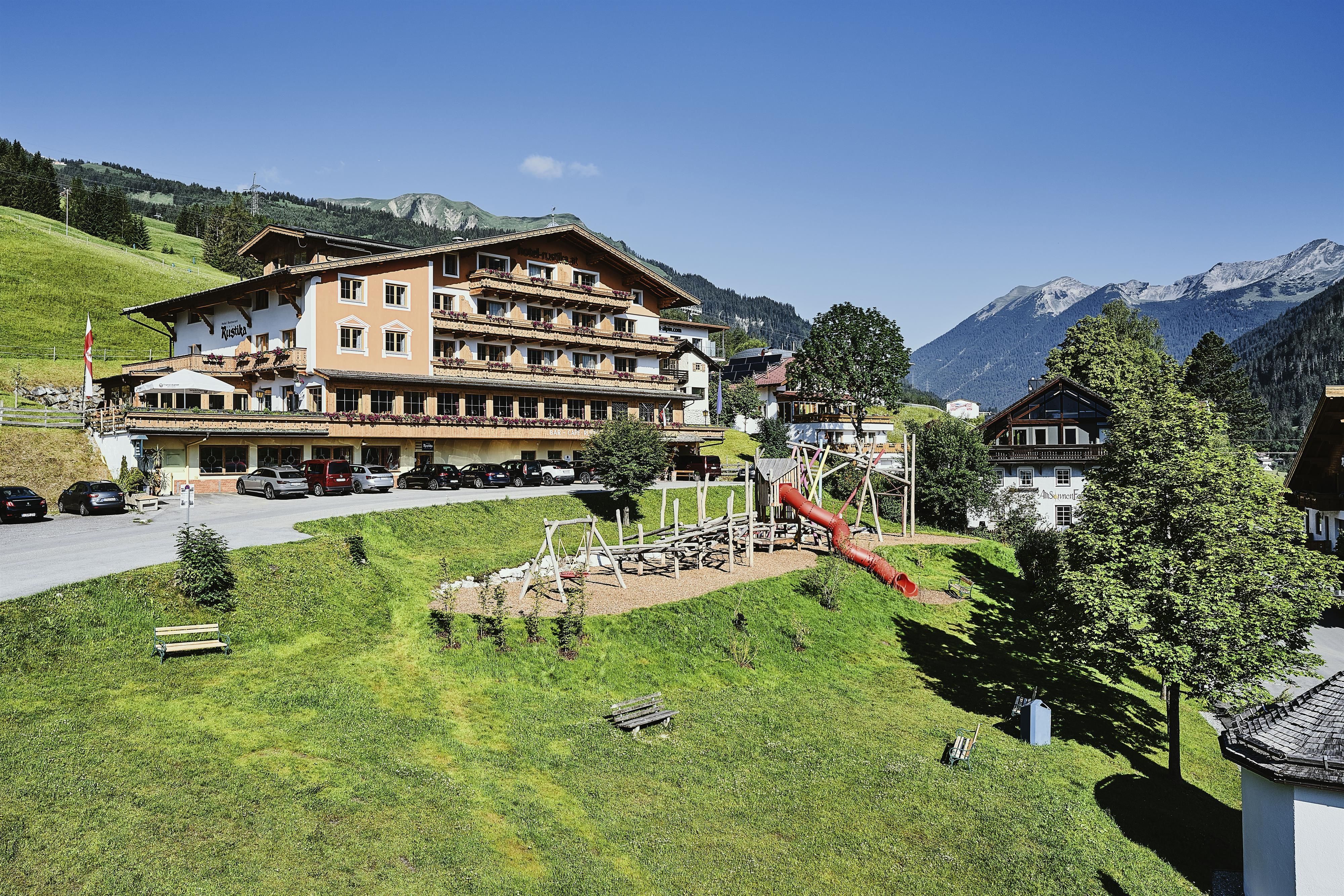 A beautiful hotel in the mountains with a playground in the foreground. The surroundings are green and wonderful with mountains in the background.