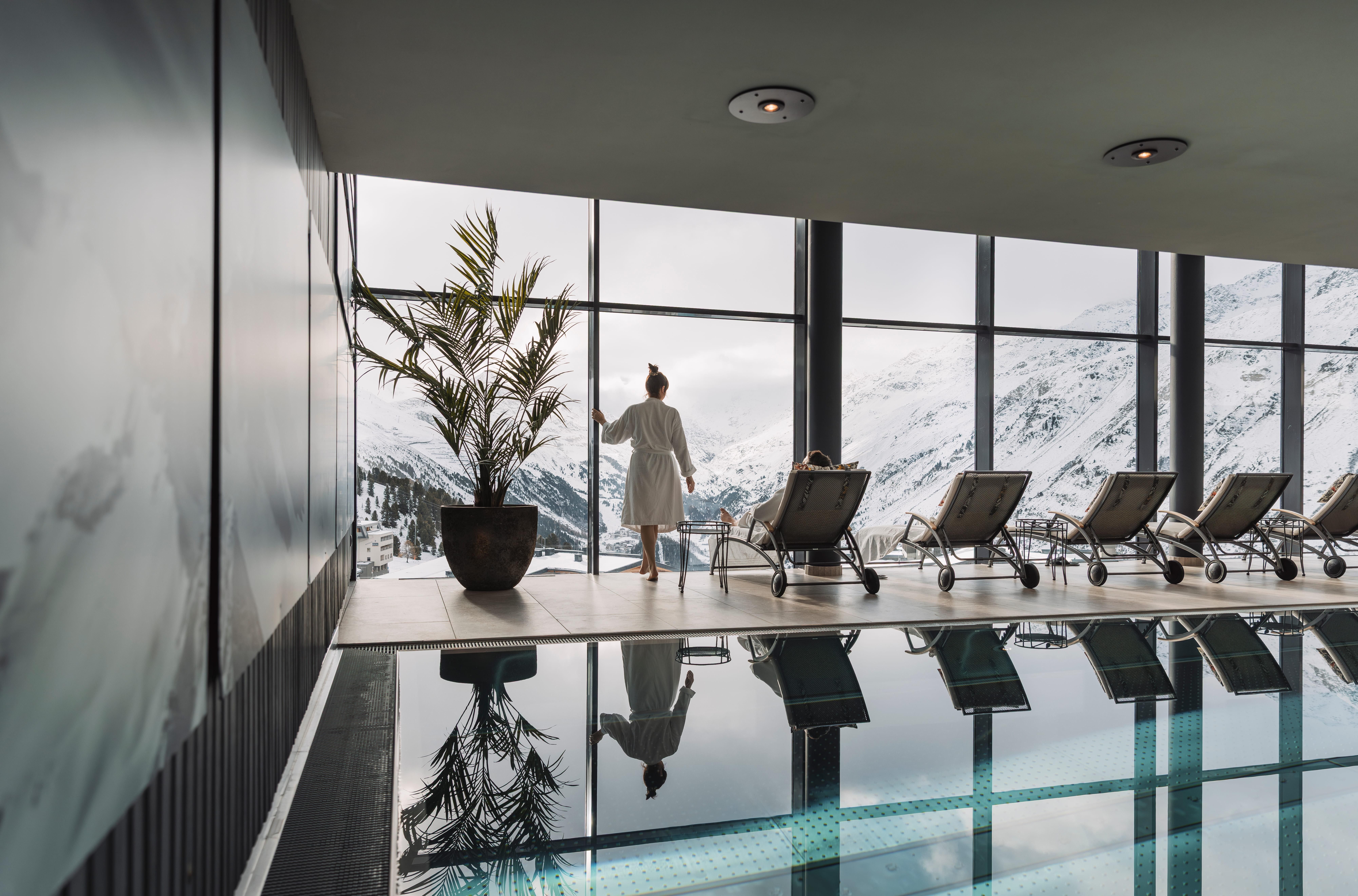 An elegant spa area with a pool and large windows overlooking snow-covered mountains. A person in a bathrobe stands relaxed and enjoys the view.
