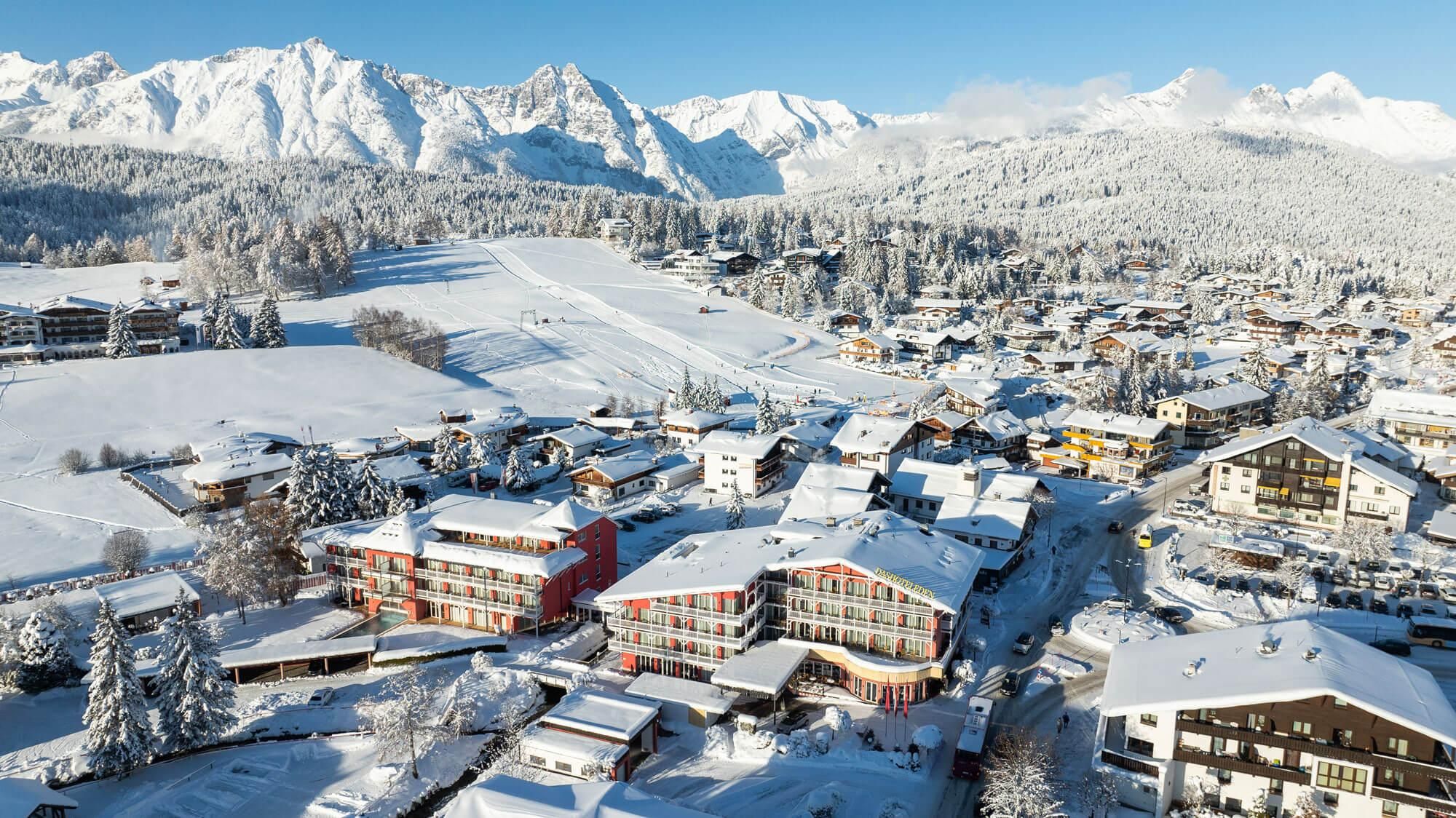 A picturesque winter landscape with snow-covered mountains and a charming village. The buildings are covered in snow, exuding a calm, wintry atmosphere.