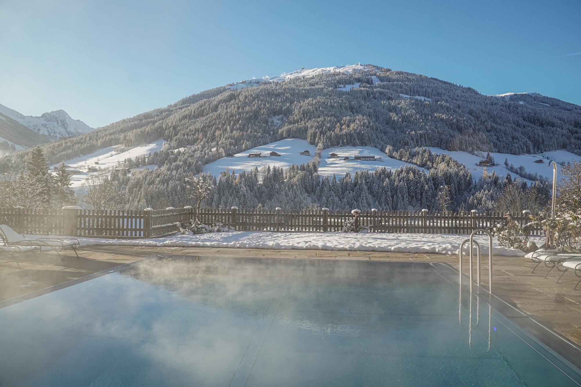 A heated pool with steaming water in front of a snow-covered mountain landscape. The sky is clear and the view is breathtaking.